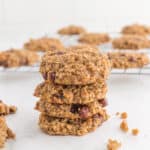 A stack of four Flourless oatmeal cookies with visible cranberries sits on a white surface, while more cookies cool on a wire rack in the background.