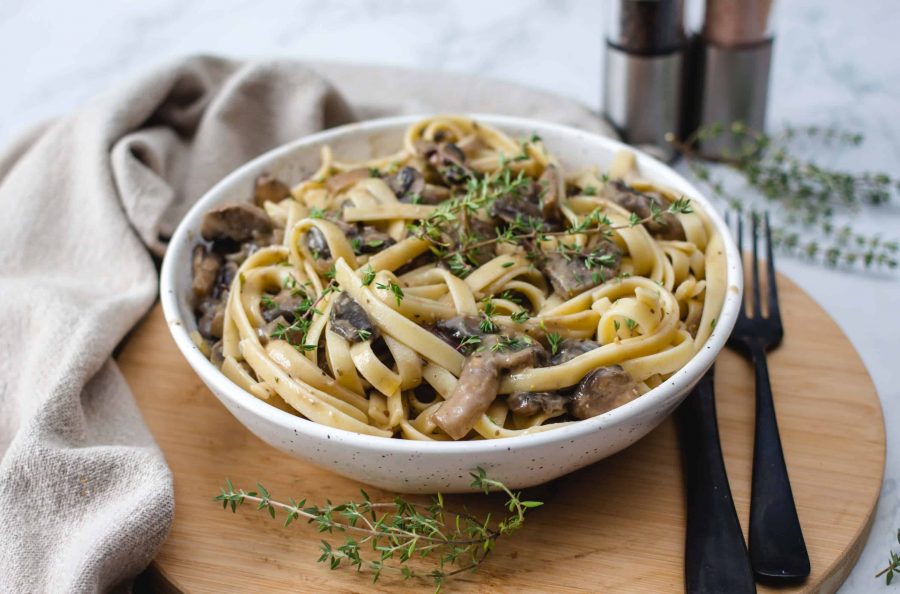 A bowl of fettuccine pasta with mushrooms Stroganoff and herbs, placed on a wooden board with a fork, knife, and napkin beside it.