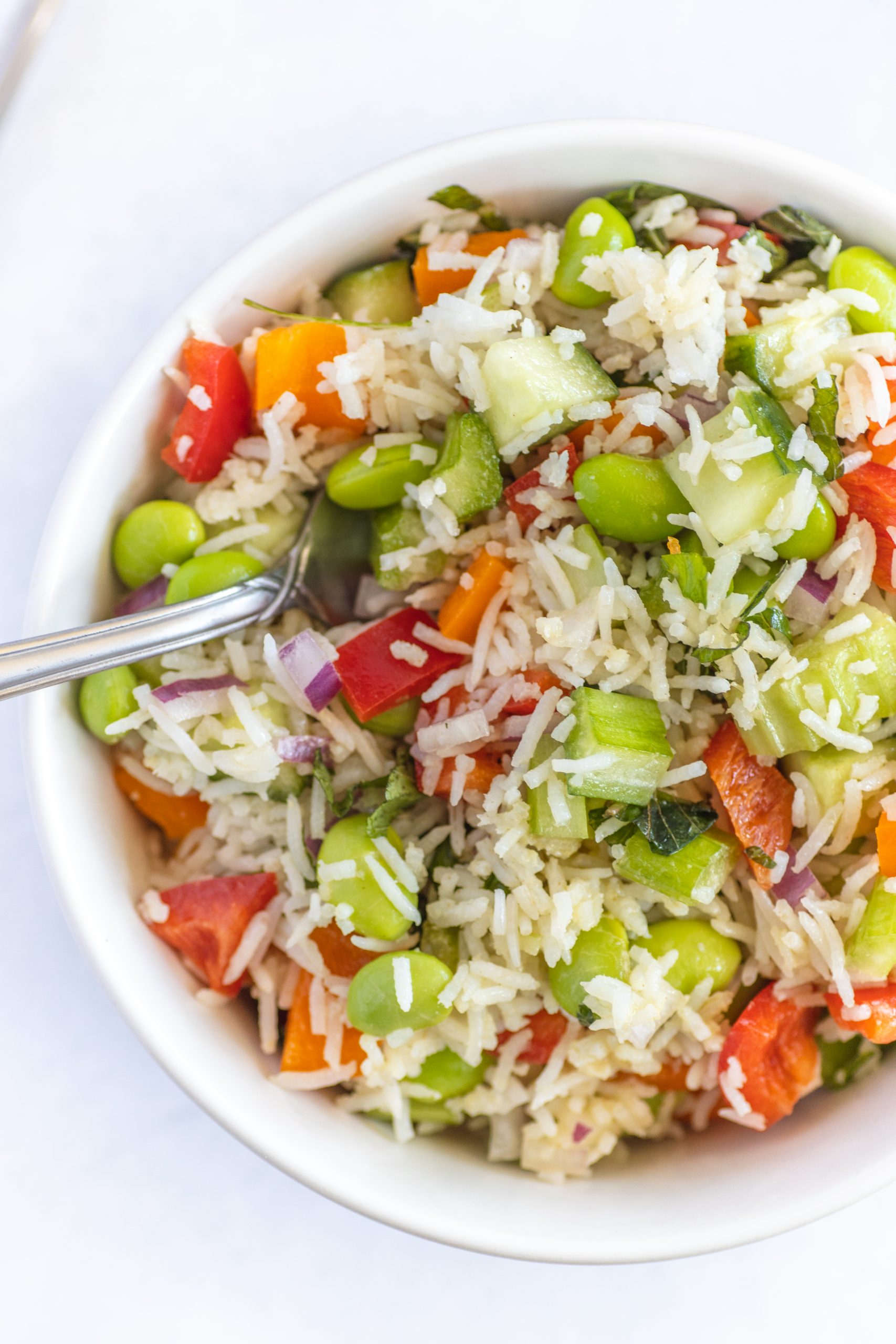A bowl of Basmati Rice Salad with Edamame, diced red bell pepper, cucumber, red onion, and fresh herbs, with a fork resting inside the bowl.