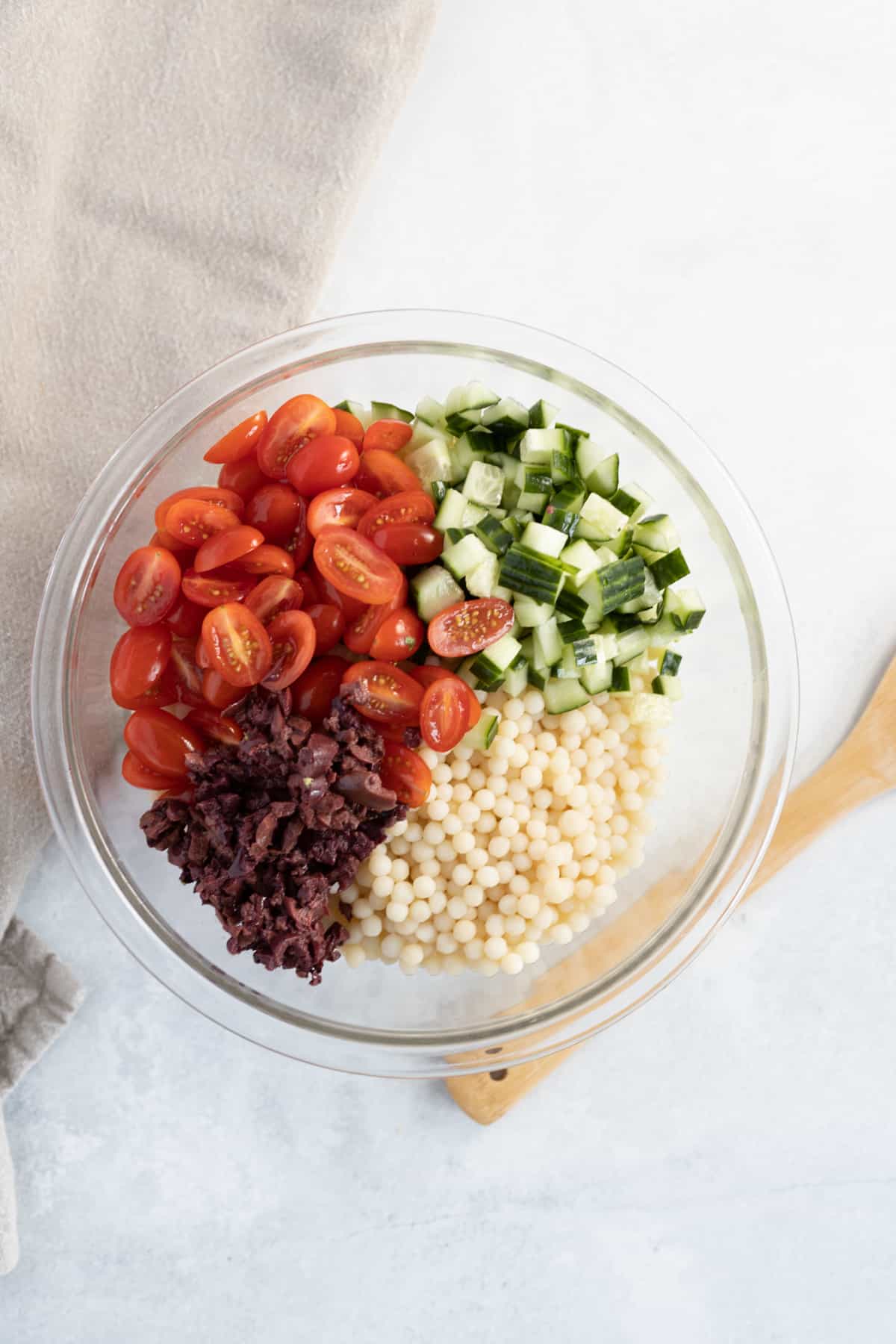 Un bol en verre contenant des tomates cerises hachées, du concombre en dés, des olives noires tranchées et du couscous perlé disposés en sections séparées pour une salade de couscous perlé vibrante.
