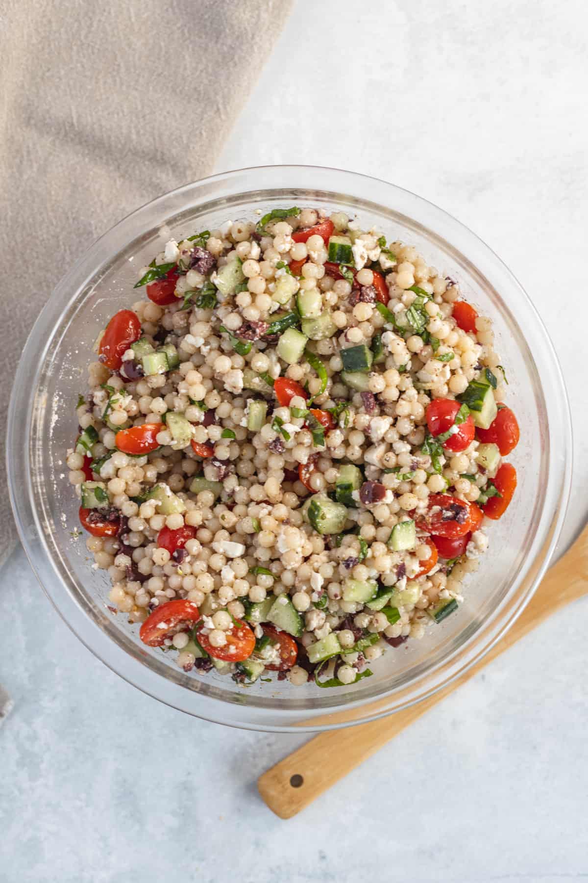 Un bol en verre rempli de salade de couscous perlé, composée de tomates cerises, de concombre, d'olives noires et d'herbes fraîches, est posé sur une surface claire à côté d'une cuillère en bois.