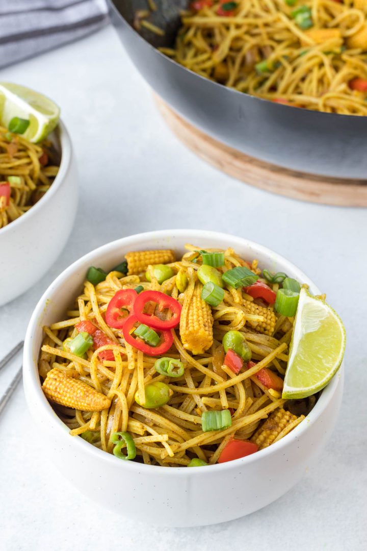 Vegetarian Singapore Noodles serve in a white bowl with wok in the background