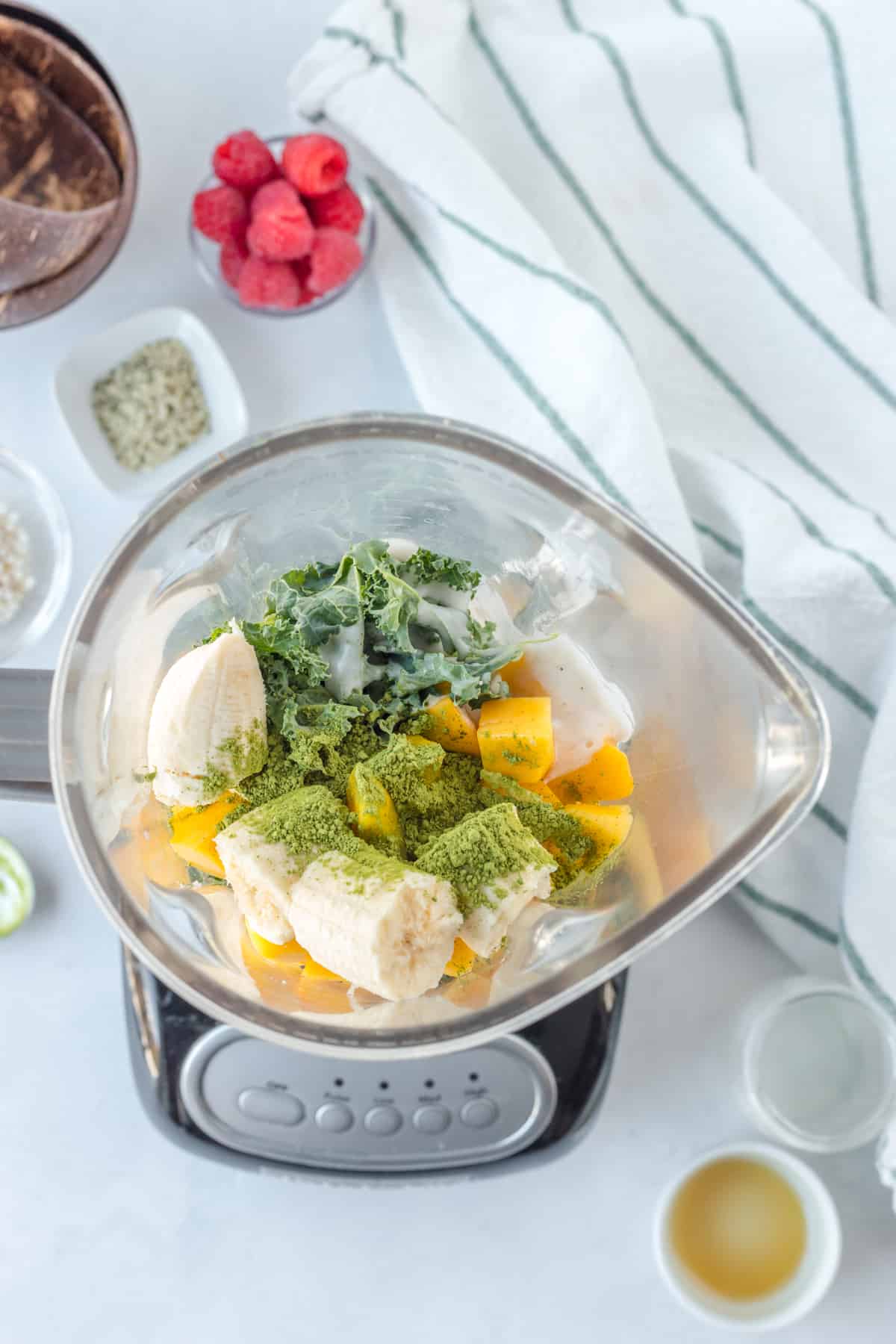 Blender filled with banana, mango, kale, and green powder for a vibrant Matcha Smoothie bowl, surrounded by small bowls of ingredients and a striped towel on a white surface.