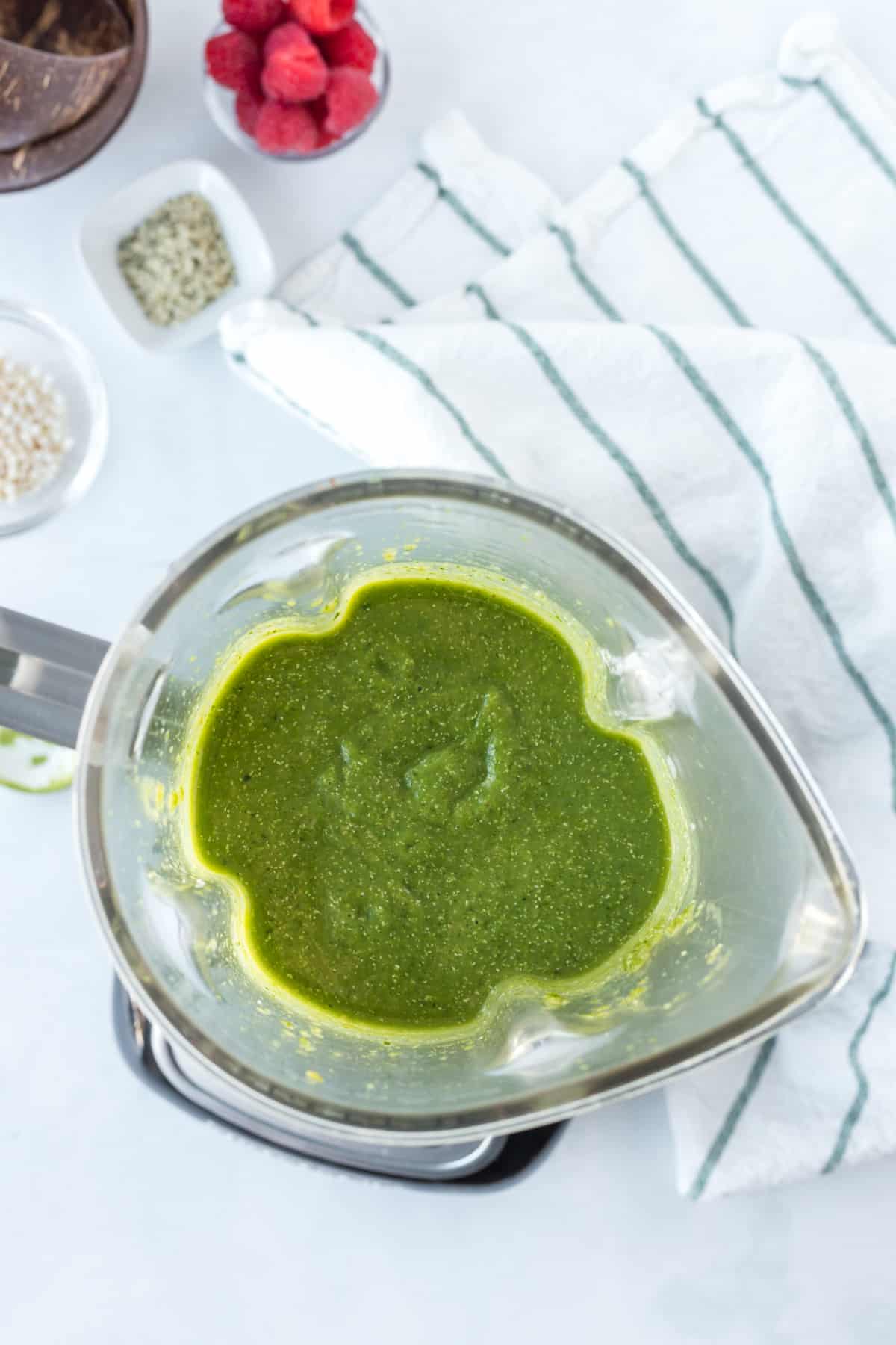 A blender filled with a vibrant matcha smoothie sits on a countertop next to a striped towel, with bowls of raspberries and seeds nearby, ready to be poured into a refreshing matcha smoothie bowl.