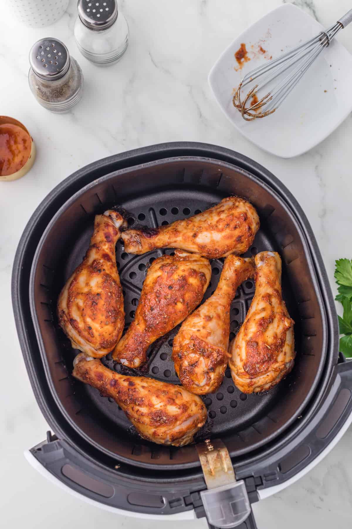 Friteuse à air avec cinq Pilons de Poulet BBQ à la friteuse à air sur un comptoir, entourés de salières et poivrières, d'un petit plat de sauce, d'un fouet usagé sur une assiette blanche et d'un brin de persil.