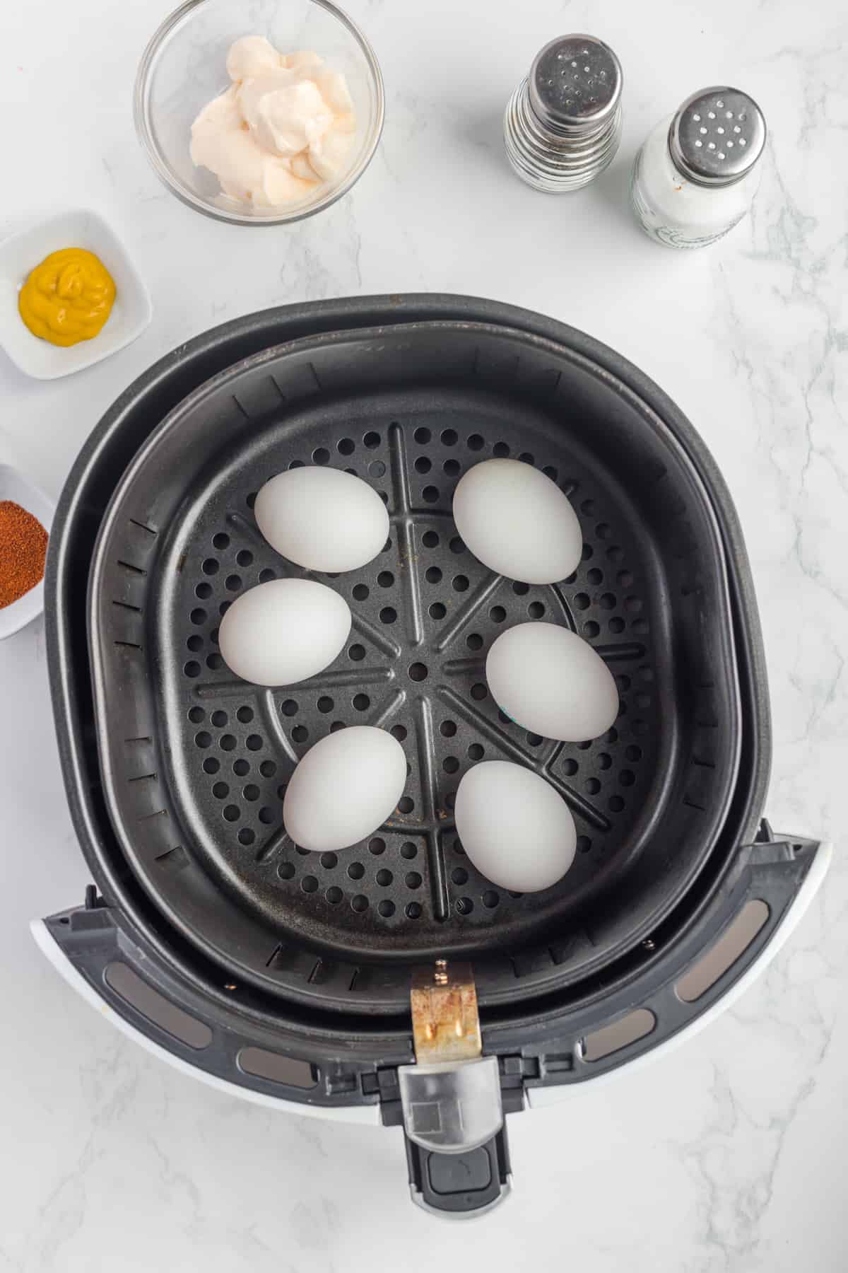 Les œufs cuits durs à la friteuse à air reposent dans un panier sur une surface en marbre, flanqué de contenants de moutarde, de mayonnaise et d'un assortiment d'épices avec des salières et des poivrières.