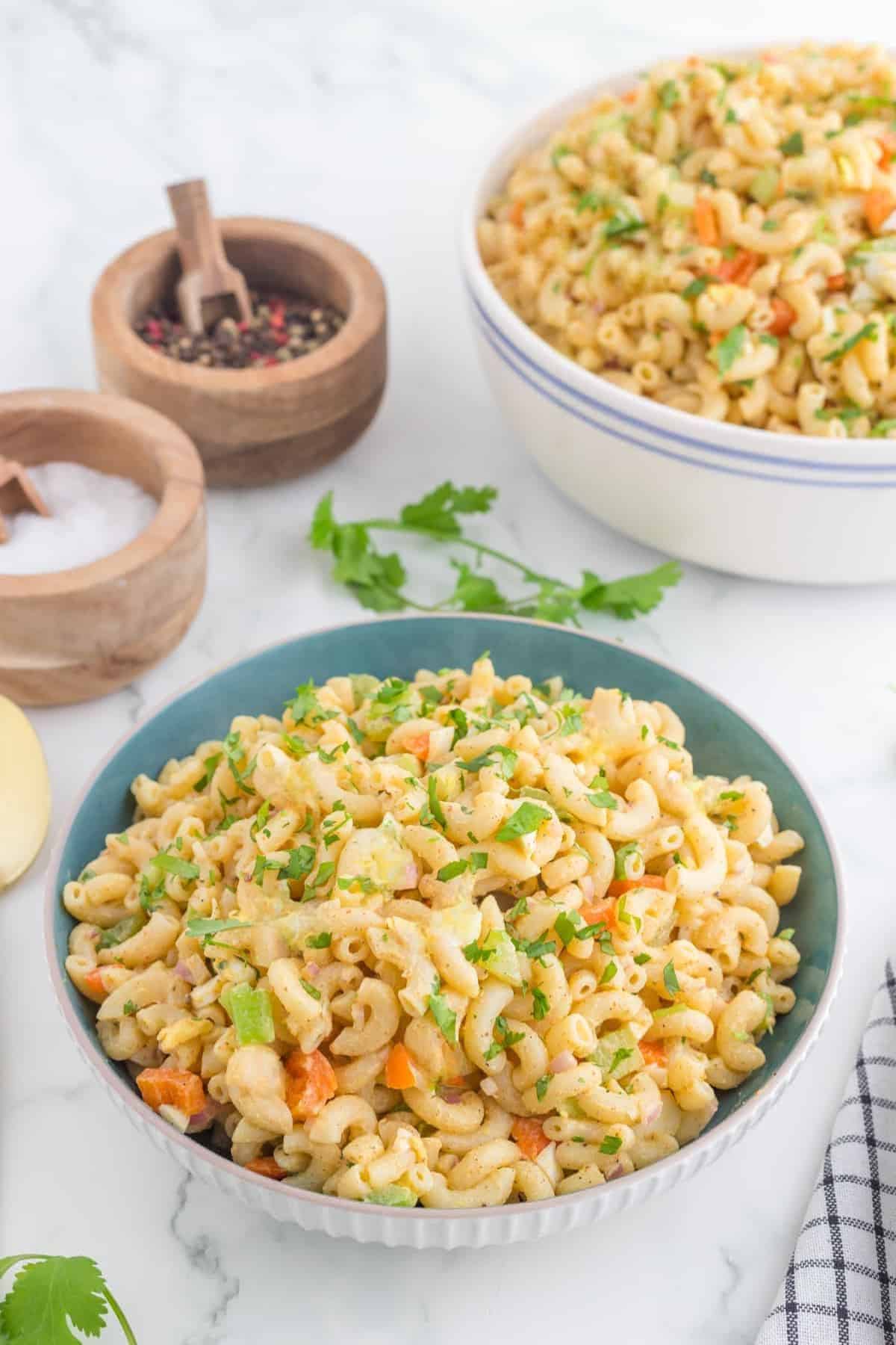Two bowls of 4 ingredients Macaroni Salad with chopped vegetables and herbs are placed on a marble surface beside wooden containers of salt and pepper.