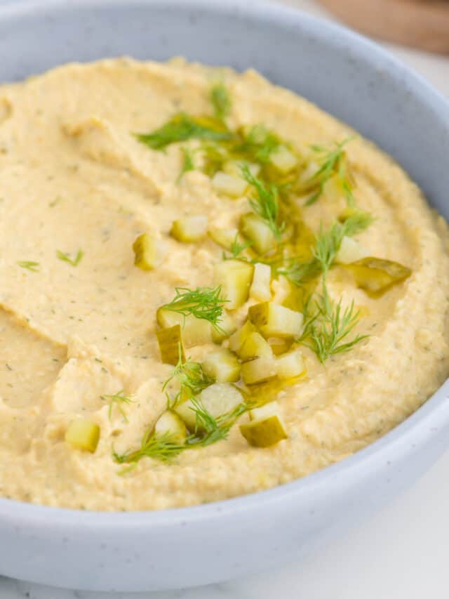 A bowl of creamy hummus topped with chopped pickles and fresh dill.