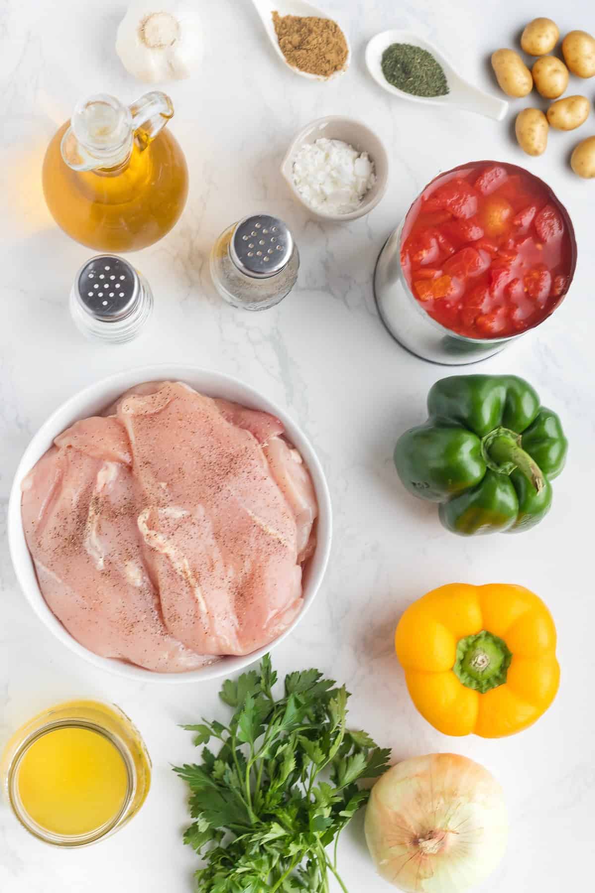 Raw chicken breasts, bell peppers, onion, parsley, canned tomatoes, potatoes, olive oil, broth, garlic, seasoning, and spices arranged on a white surface-perfect ingredients for a flavorful Chicken Goulash.