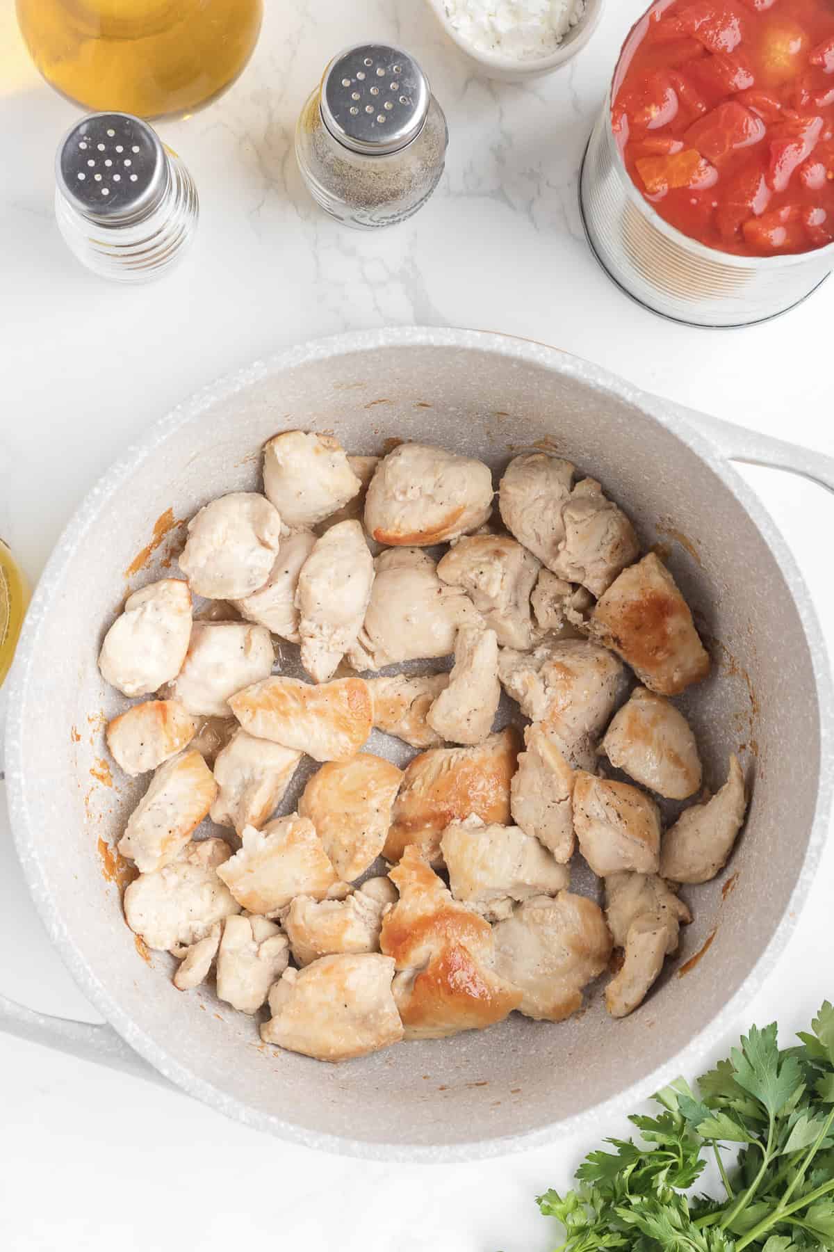 Chunks of cooked chicken in a pot, surrounded by canned tomatoes, chopped onions, seasonings, olive oil, and fresh parsley on a white countertop-perfect for making hearty Chicken Goulash.