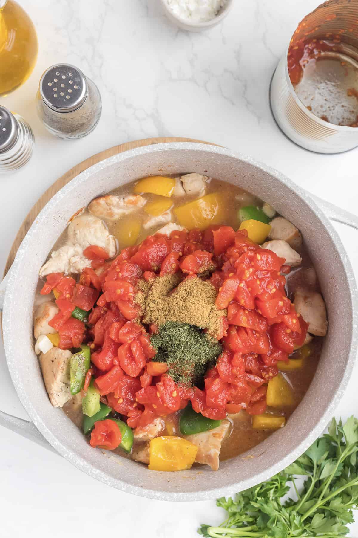 A pot with chopped chicken, bell peppers, diced tomatoes, and spices ready to be cooked for a flavorful Chicken Goulash, surrounded by seasonings, oil, and parsley on a white countertop.