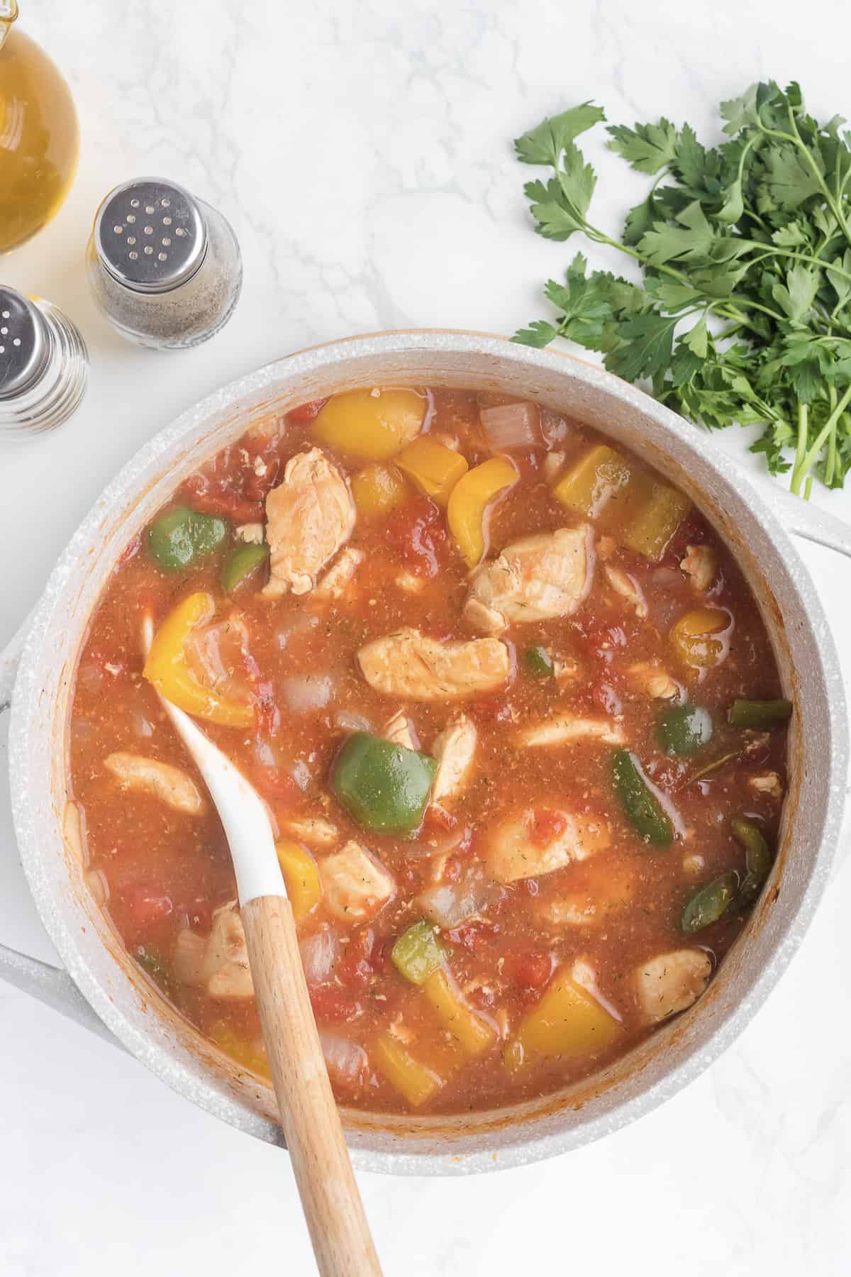 A pot of Cajun Chicken Stew with bell peppers and tomatoes sits on a white surface beside a wooden spoon, fresh parsley, salt and pepper shakers, and a bottle of oil.