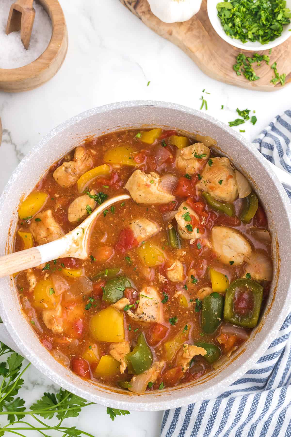 A pot of Cajun Chicken stew with diced chicken, bell peppers, onions, potatoes and tomatoes in a red broth, with a wooden spoon resting inside. Fresh herbs are sprinkled on top.