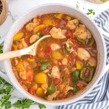 A pot ofCajun chicken Stew with chunks of chicken, bell peppers, onions, and tomatoes sits ready to serve, accompanied by a white ladle and a striped cloth nearby.