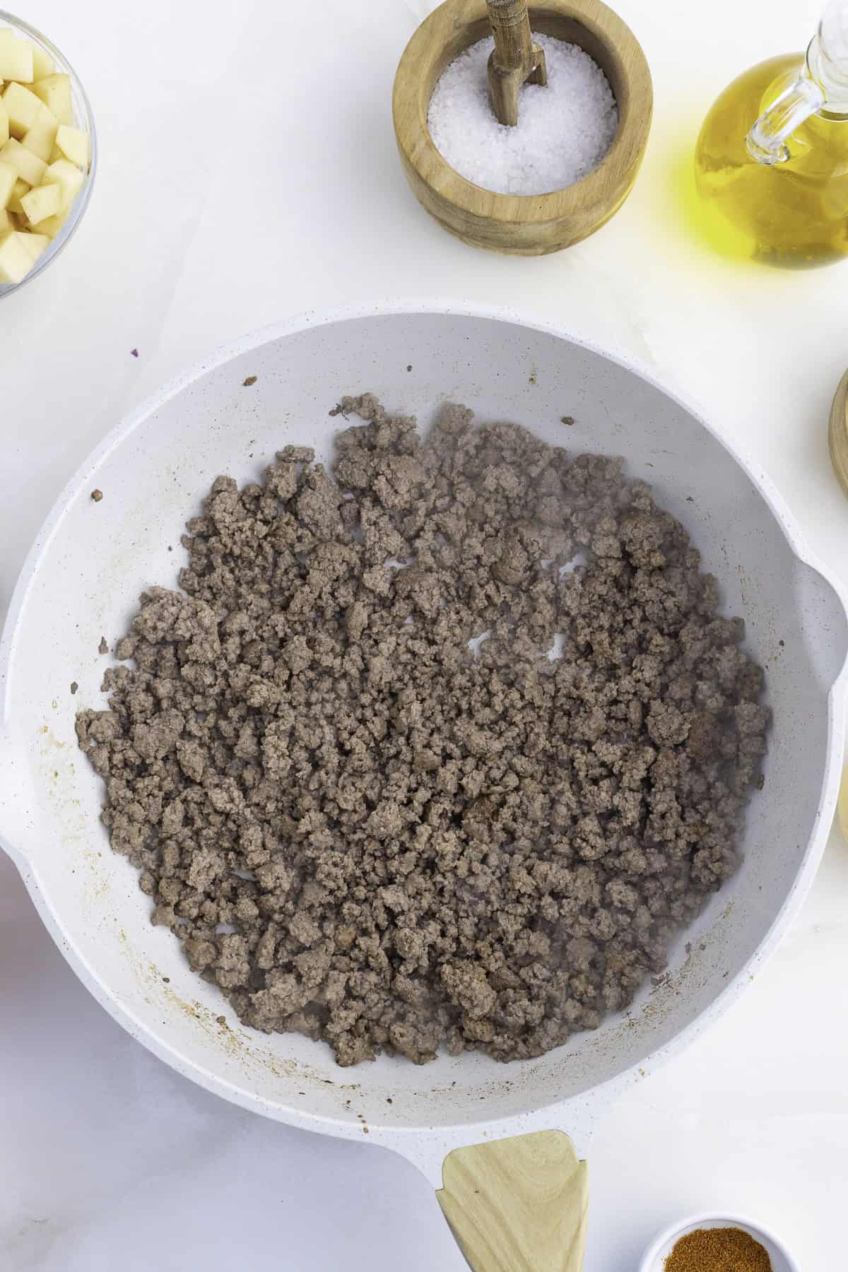 Ground beef cooking in a white skillet on a marble surface, with bowls of salt, diced potatoes, and a glass bottle of olive oil nearby.