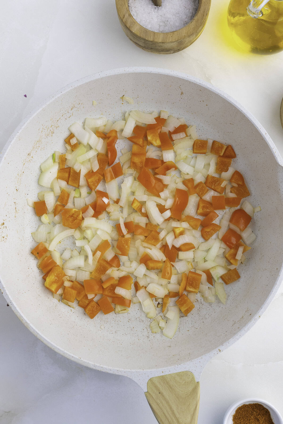 Chopped onions and orange bell peppers cooking in a white frying pan, surrounded by a container of salt, a bottle of oil, and a small bowl of spice.