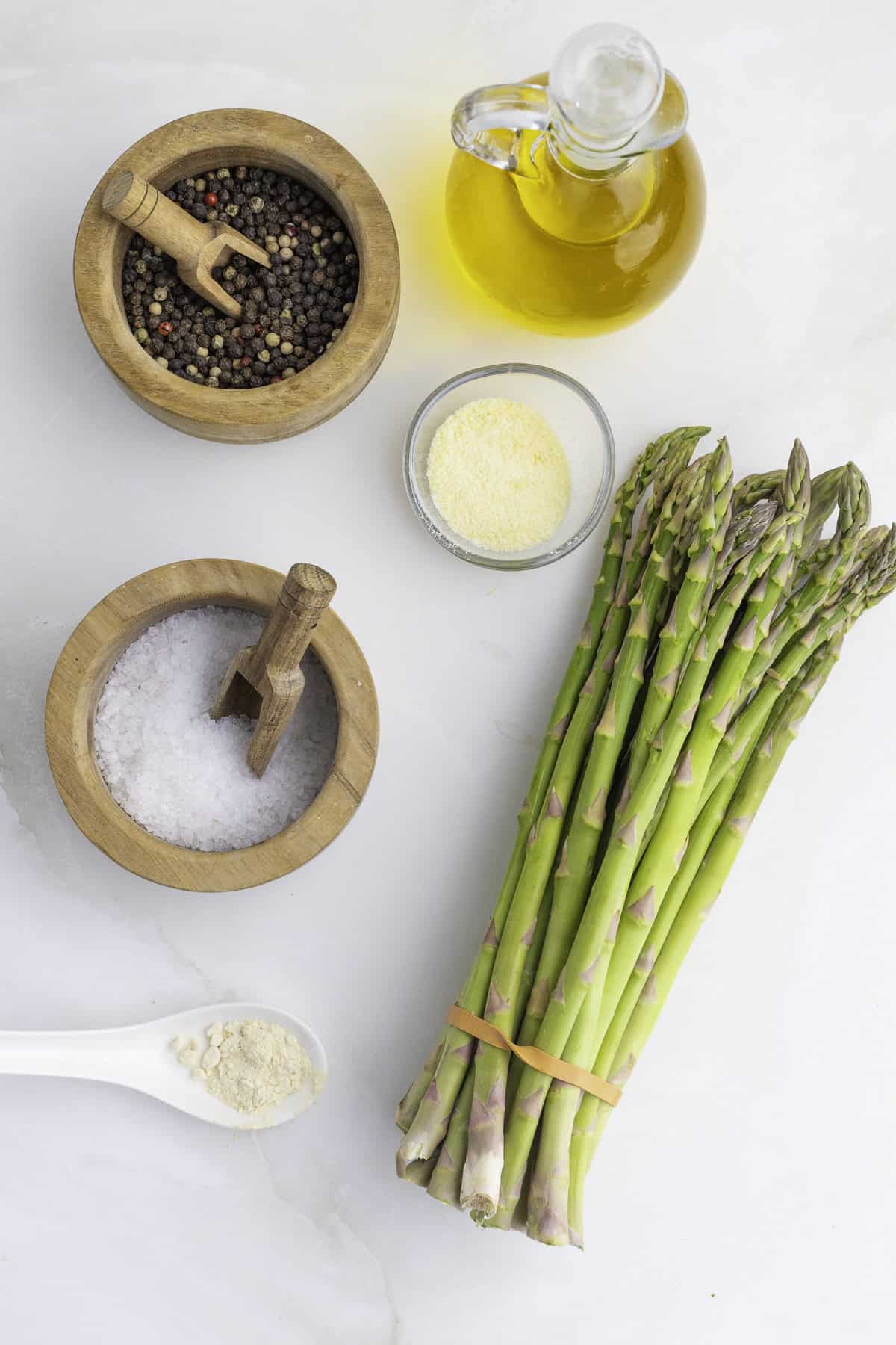 Asparagus, olive oil, black peppercorns, sea salt, grated cheese, and garlic powder on a white surface.