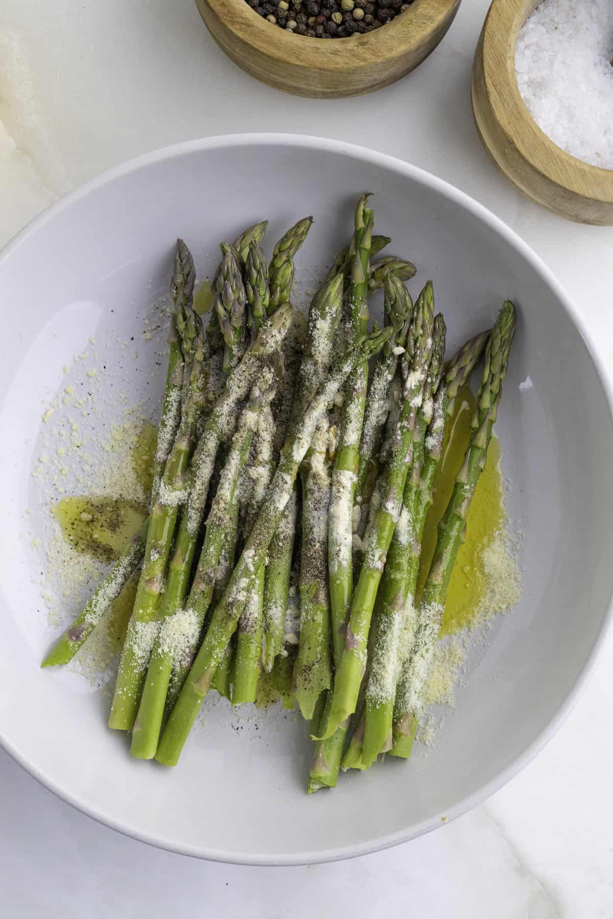 A bowl of raw asparagus spears topped with grated cheese, olive oil, and black pepper, next to bowls of salt and pepper.