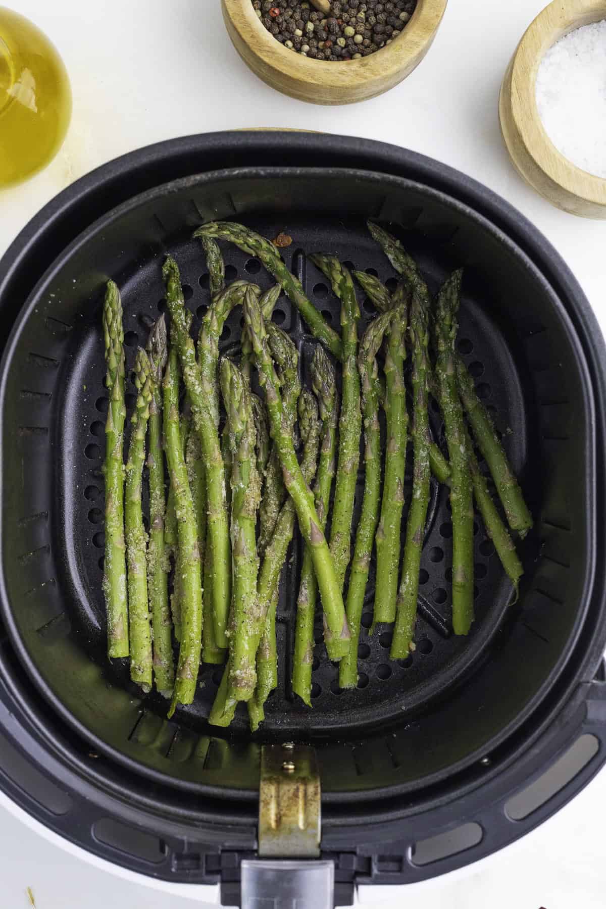 Asparagus spears in an air fryer basket, surrounded by bowls of peppercorns, salt, and a bottle of olive oil.