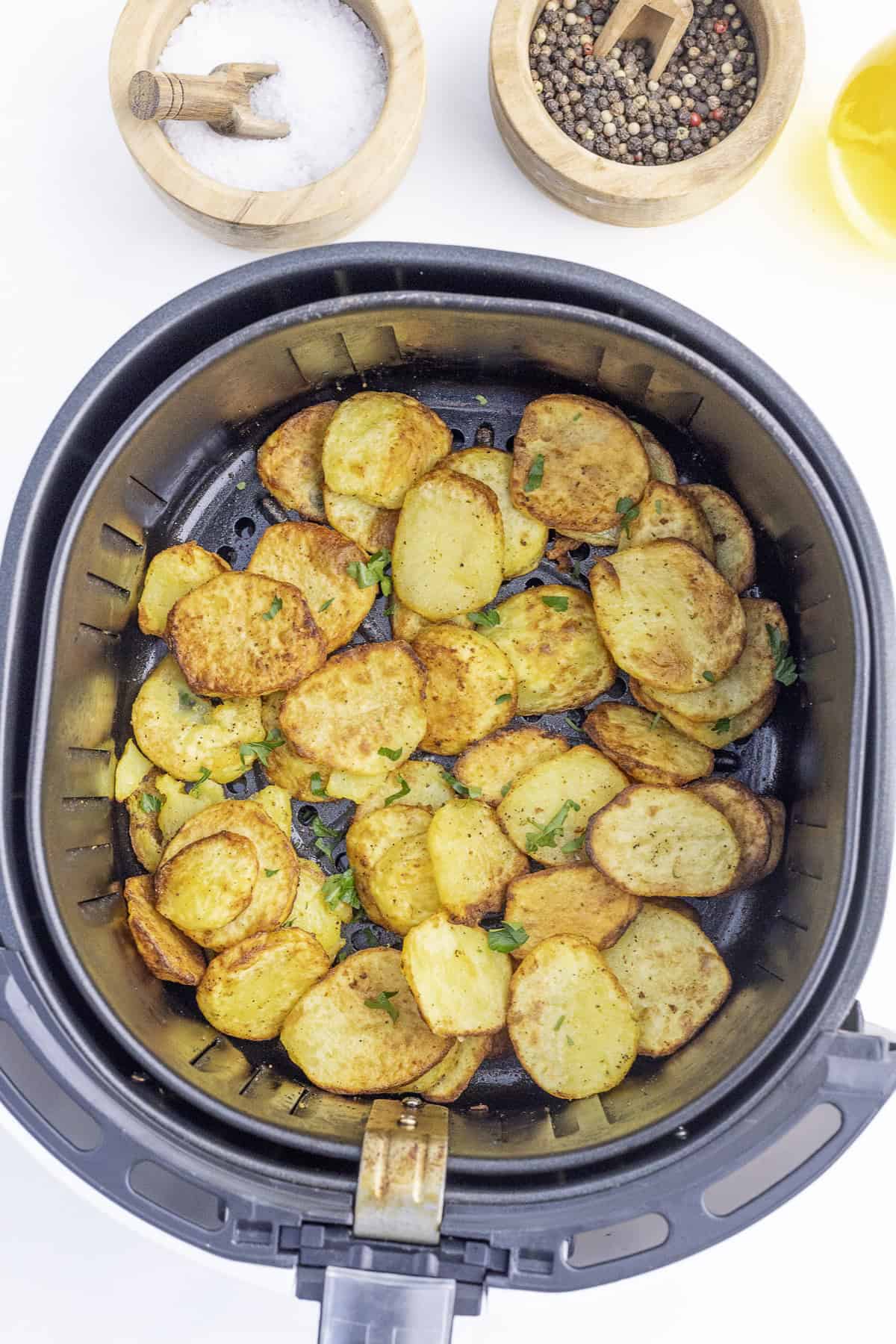 Sliced, seasoned potatoes are cooked in an air fryer basket. Nearby are small bowls of salt and pepper.