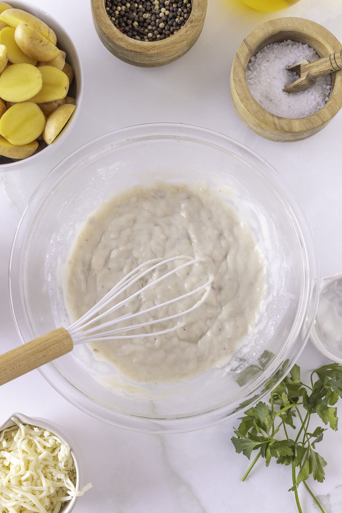 A bowl of white sauce with a whisk, surrounded by potatoes, grated cheese, parsley, salt, pepper, and olive oil on a marble surface.