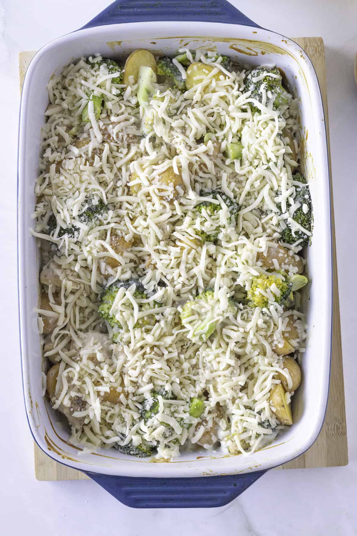 A casserole dish filled with sliced potatoes, broccoli, and topped with shredded cheese, ready for baking.