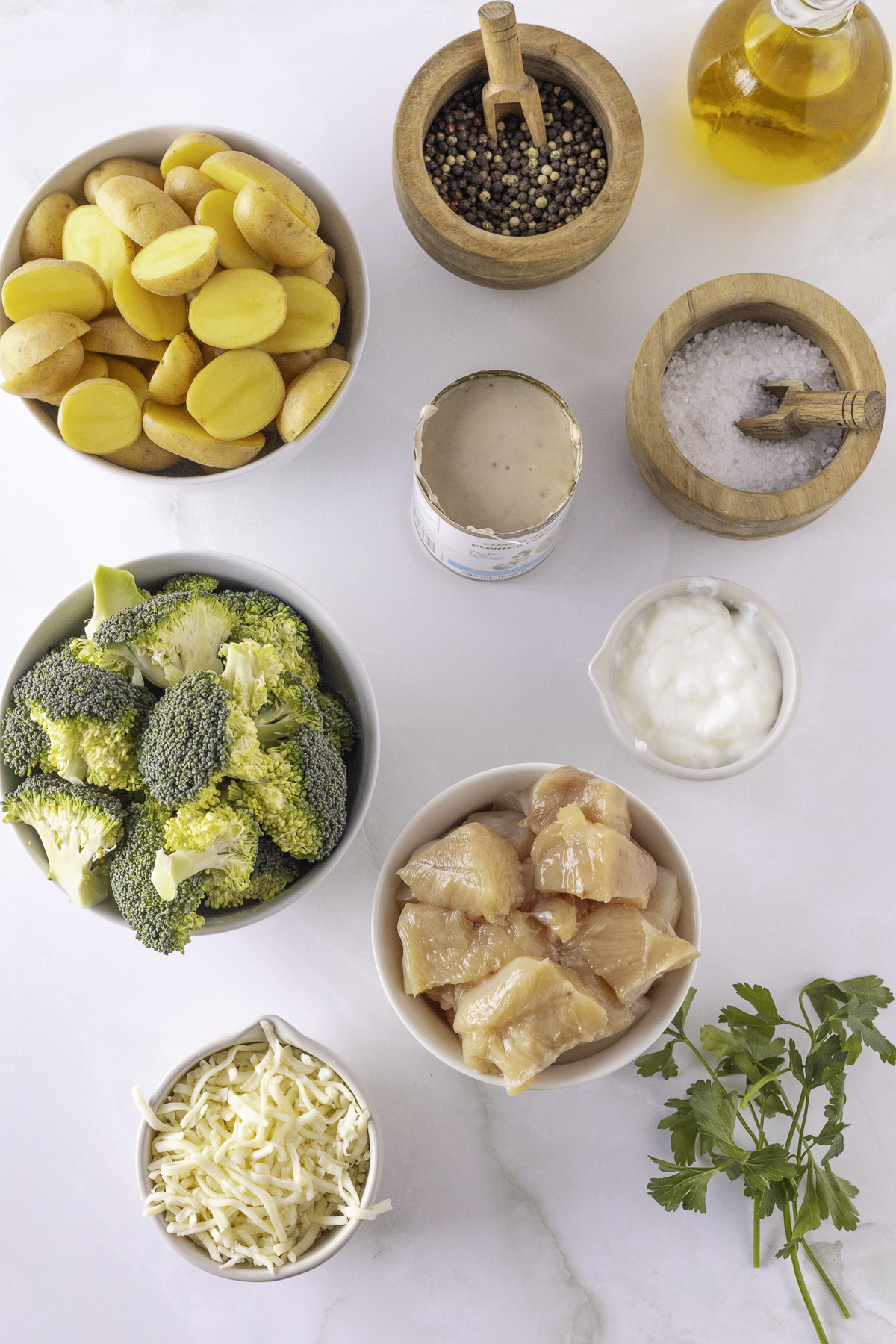 Ingredients on a table: sliced potatoes, broccoli, raw chicken chunks, shredded cheese, yogurt, pepper, salt, olive oil, parsley, and sauce in a can.