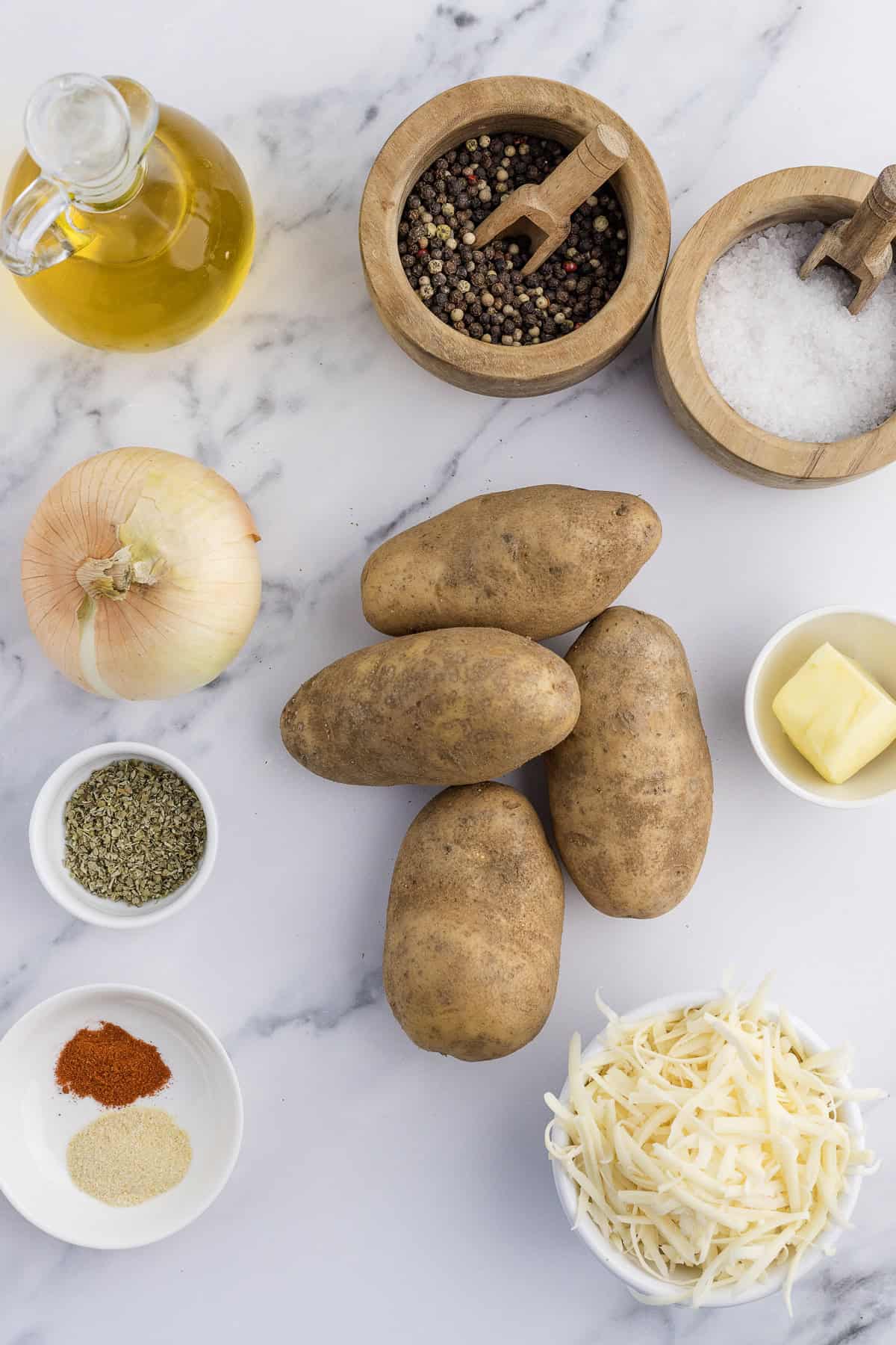 Potatoes, cheese, onion, olive oil, spices, butter, salt, and peppercorns are arranged on a marble surface.