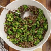 A white pan filled with cooked ground beef and broccoli florets, garnished with sesame seeds, and a serving spoon resting inside.