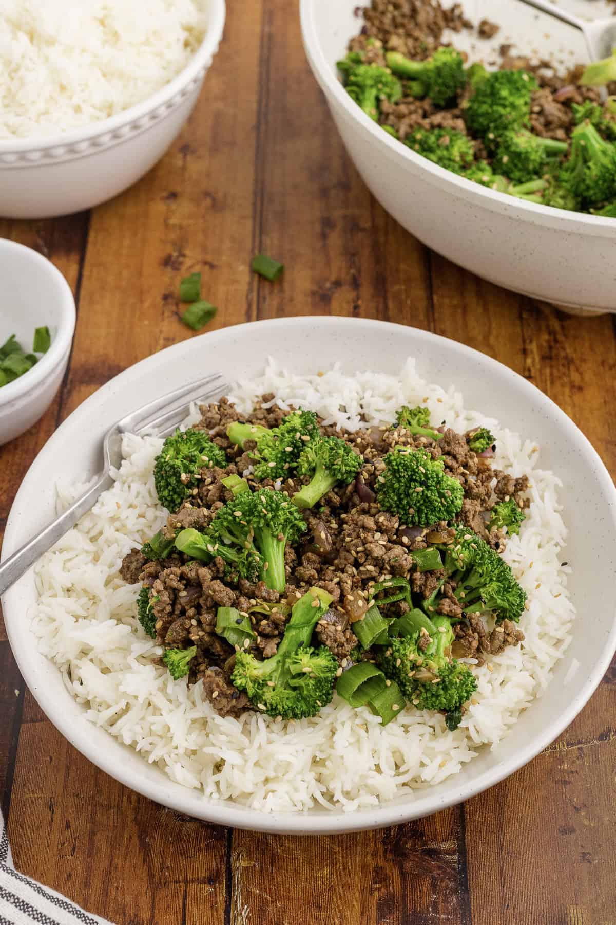 A dish of white rice topped with cooked ground beef and broccoli served on a wooden table. A fork rests on the plate, and additional bowls of rice and beef with broccoli are nearby.