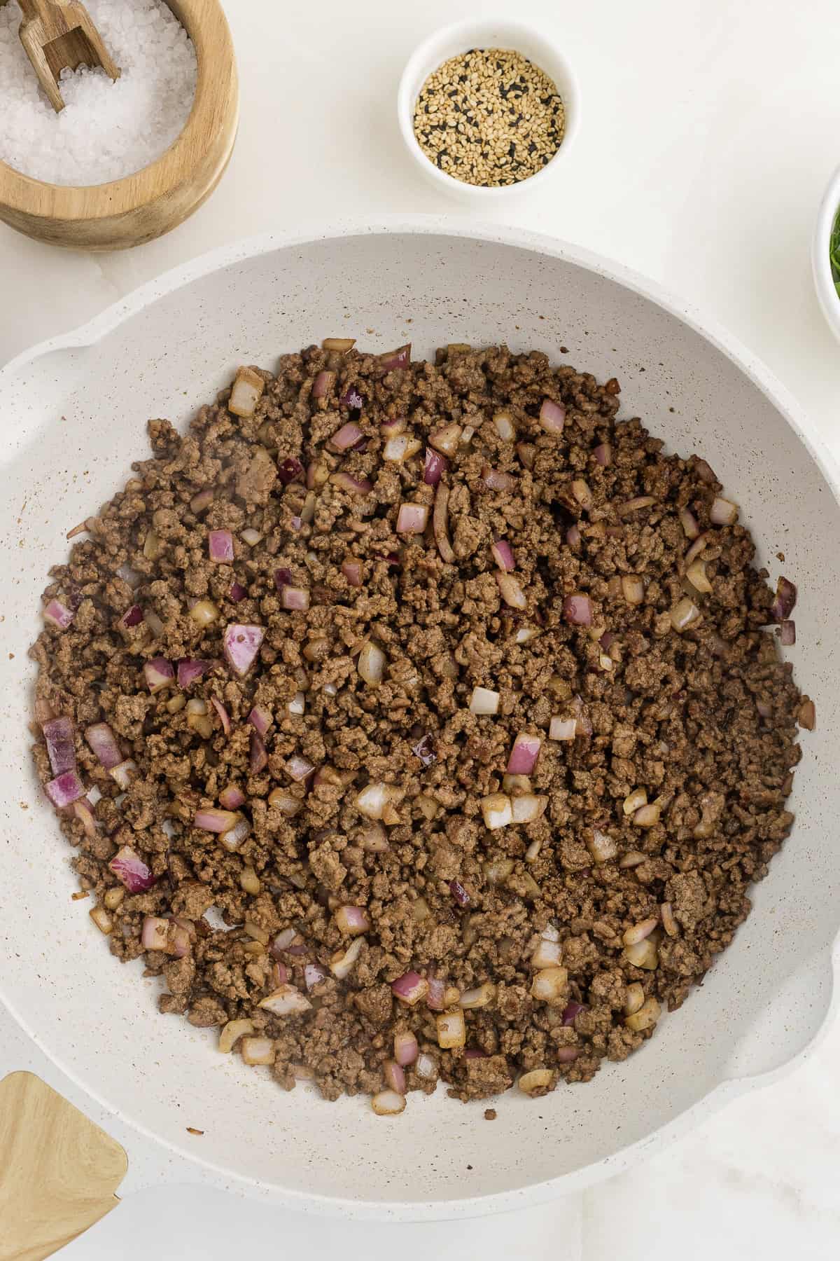 A white pan containing cooked ground beef mixed with chopped red onions. Adjacent are bowls with salt, seeds, and green herbs.