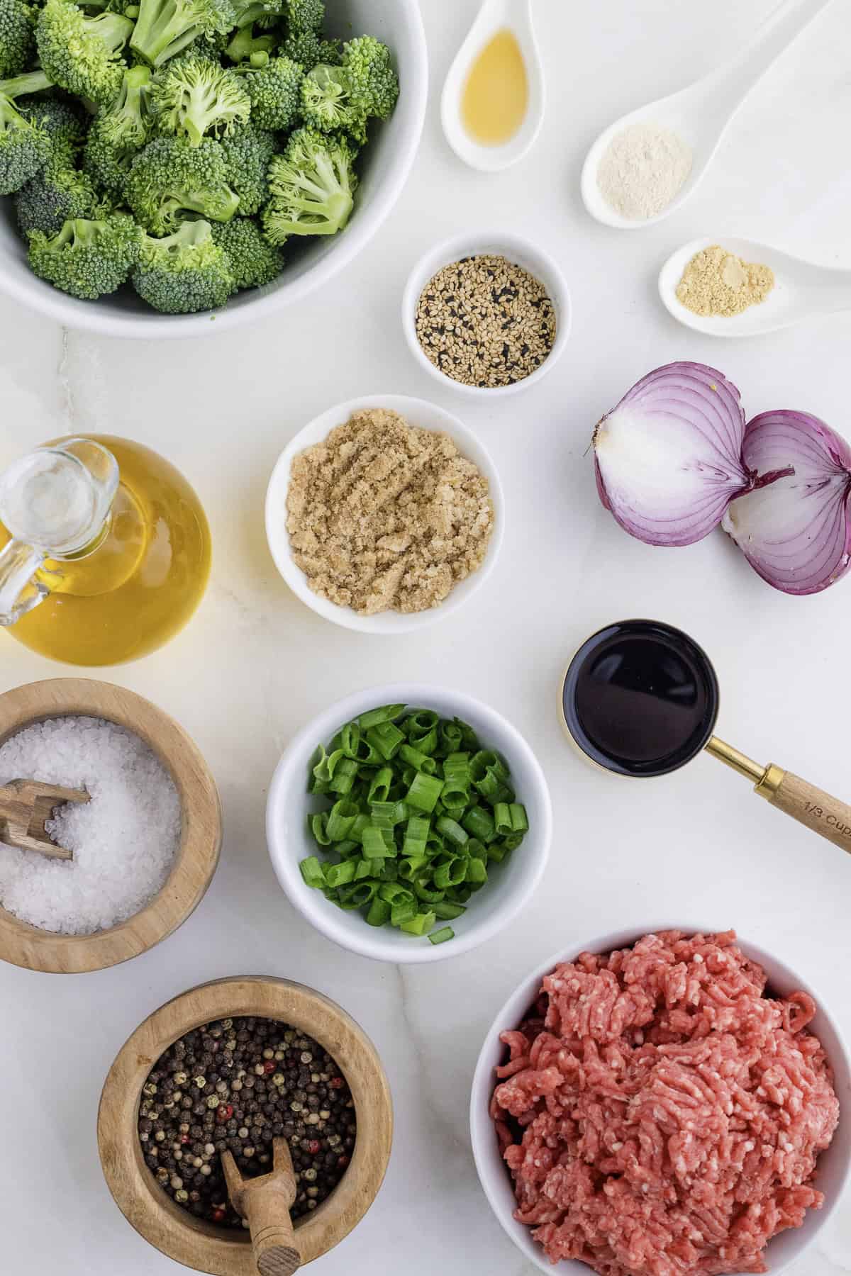 Assorted ingredients on a countertop including broccoli, onion, ground meat, spices, chopped green onions, brown sugar, soy sauce, olive oil, and seasoning.