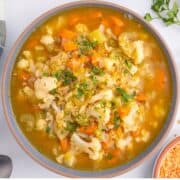 A bowl of vegetable soup with cauliflower, carrots, celery, and parsley garnish. A spoon and scattered lentils are nearby on a white surface.