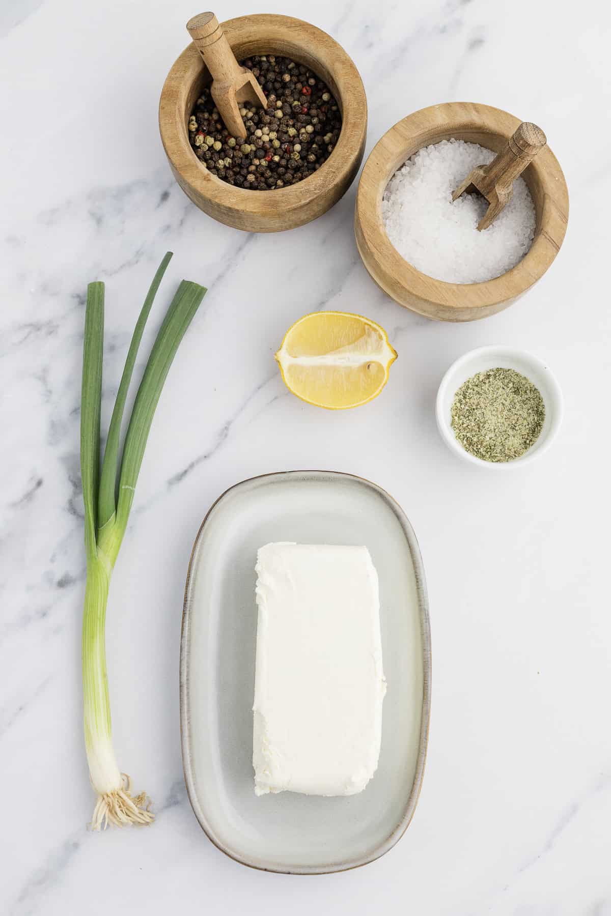 A slab of cream cheese on a plate, surrounded by green onions, a lemon wedge, small wooden bowls of peppercorns and salt, and a bowl of mixed herbs on a marble surface.