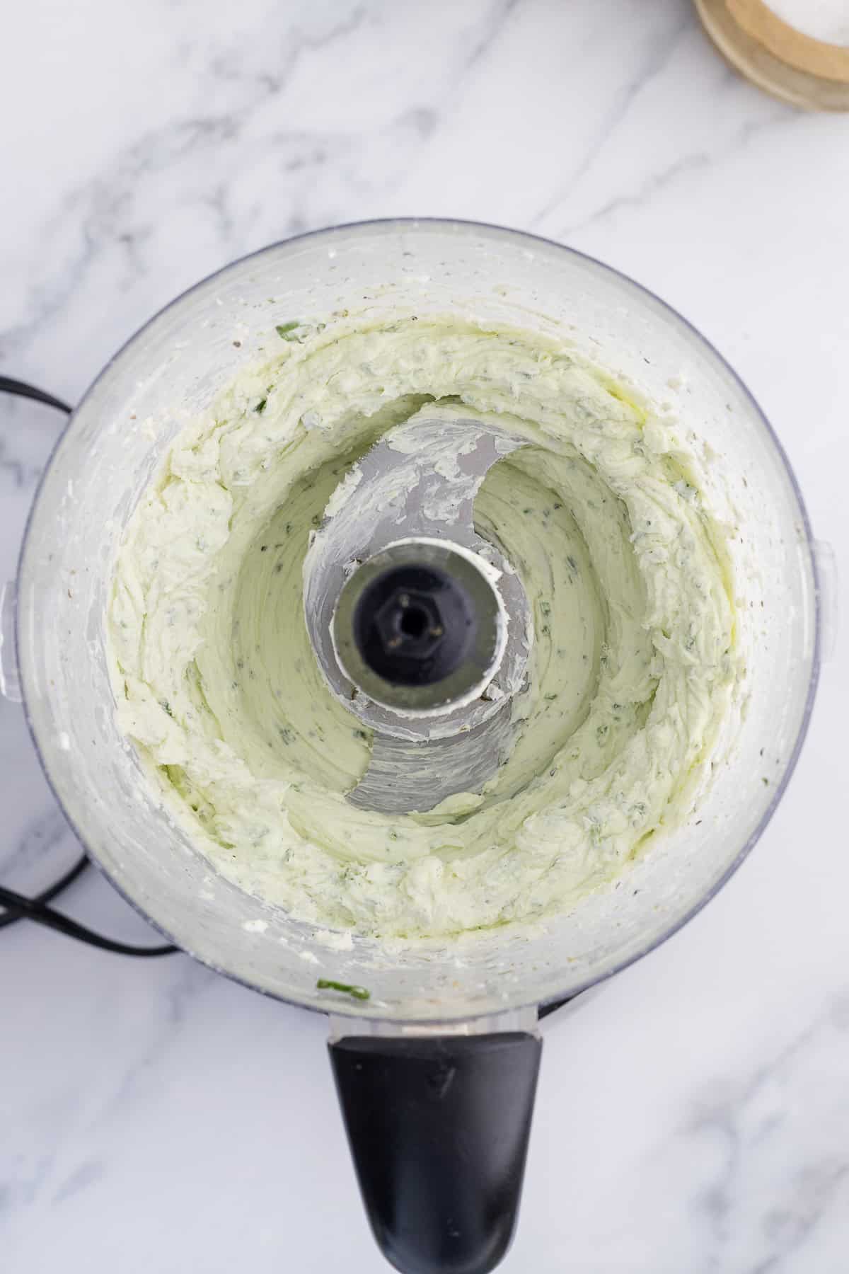 Top view of a food processor with creamy herb mixture, featuring a pale green color and visible herb specks, placed on a marble countertop.