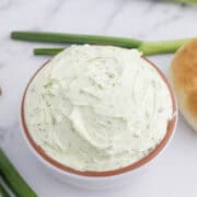 A bowl of creamy green onion dip on a marble surface, surrounded by green onions and a bun.