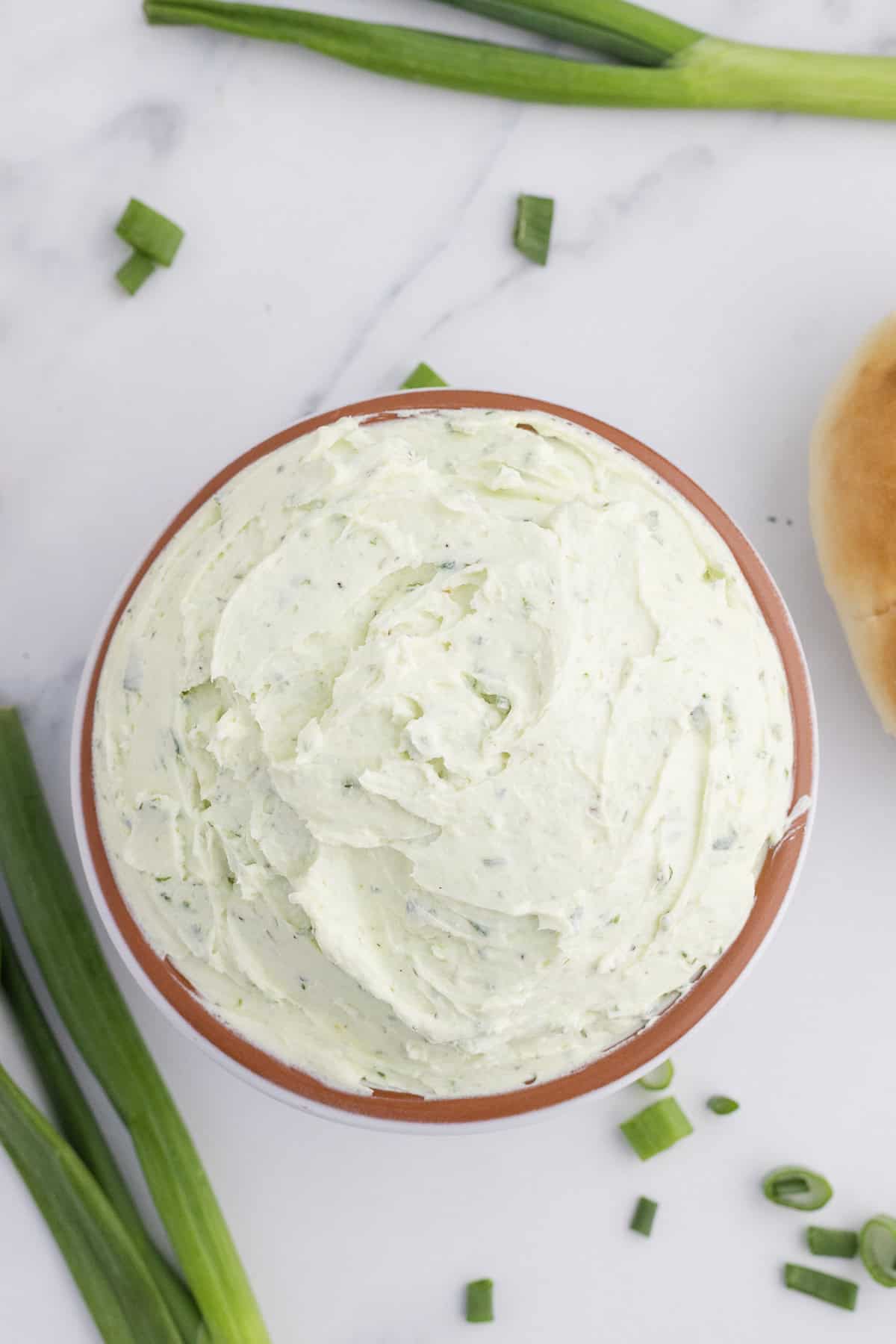 A bowl of creamy, whipped chive butter on a marble surface, surrounded by green onions and a bread roll.