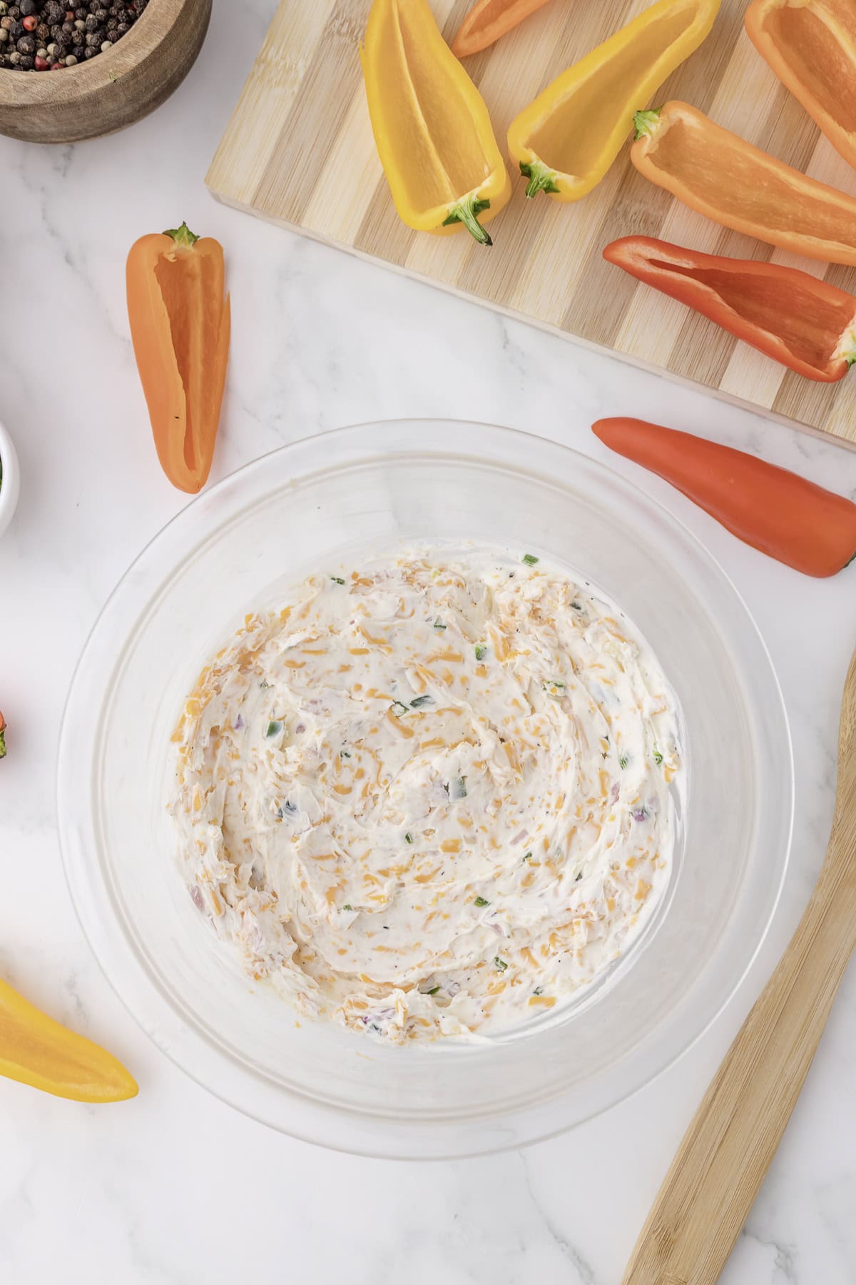 Bowl of creamy dip with herbs and cheese on a marble surface, surrounded by sliced colorful bell peppers on a wooden board.
