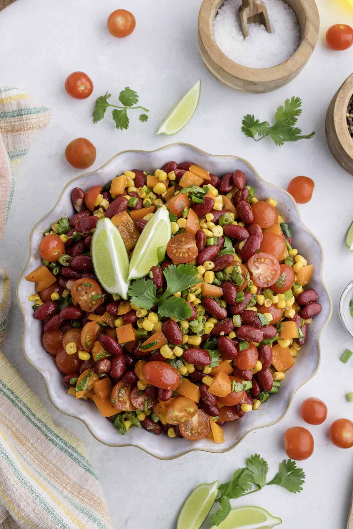 A bowl of bean salad with kidney beans, corn, cherry tomatoes, bell peppers, and cilantro, garnished with lime wedges. Surrounding are lime slices, tomatoes, and small bowls on a light surface.