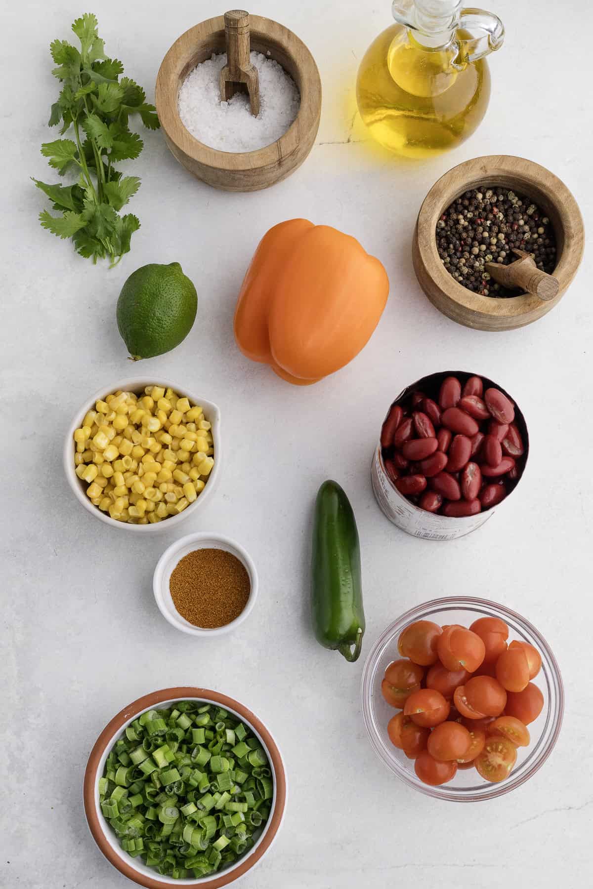 Cherry tomatoes, kidney beans, corn, jalapeño, bell pepper, lime, scallions, cilantro, oil, salt, pepper, and spices are arranged on a white surface.