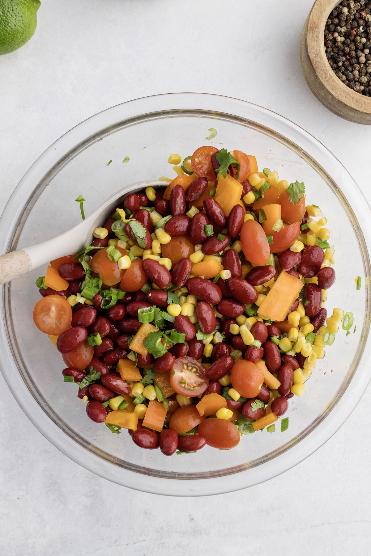 A clear glass bowl filled with a salad of red kidney beans, corn, cherry tomatoes, cubed cheese, and chopped herbs, with a wooden spoon inside. A dish with pepper is nearby.