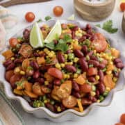 A bowl of kidney bean salad with corn, cherry tomatoes, bell peppers, and garnished with lime wedges and cilantro.