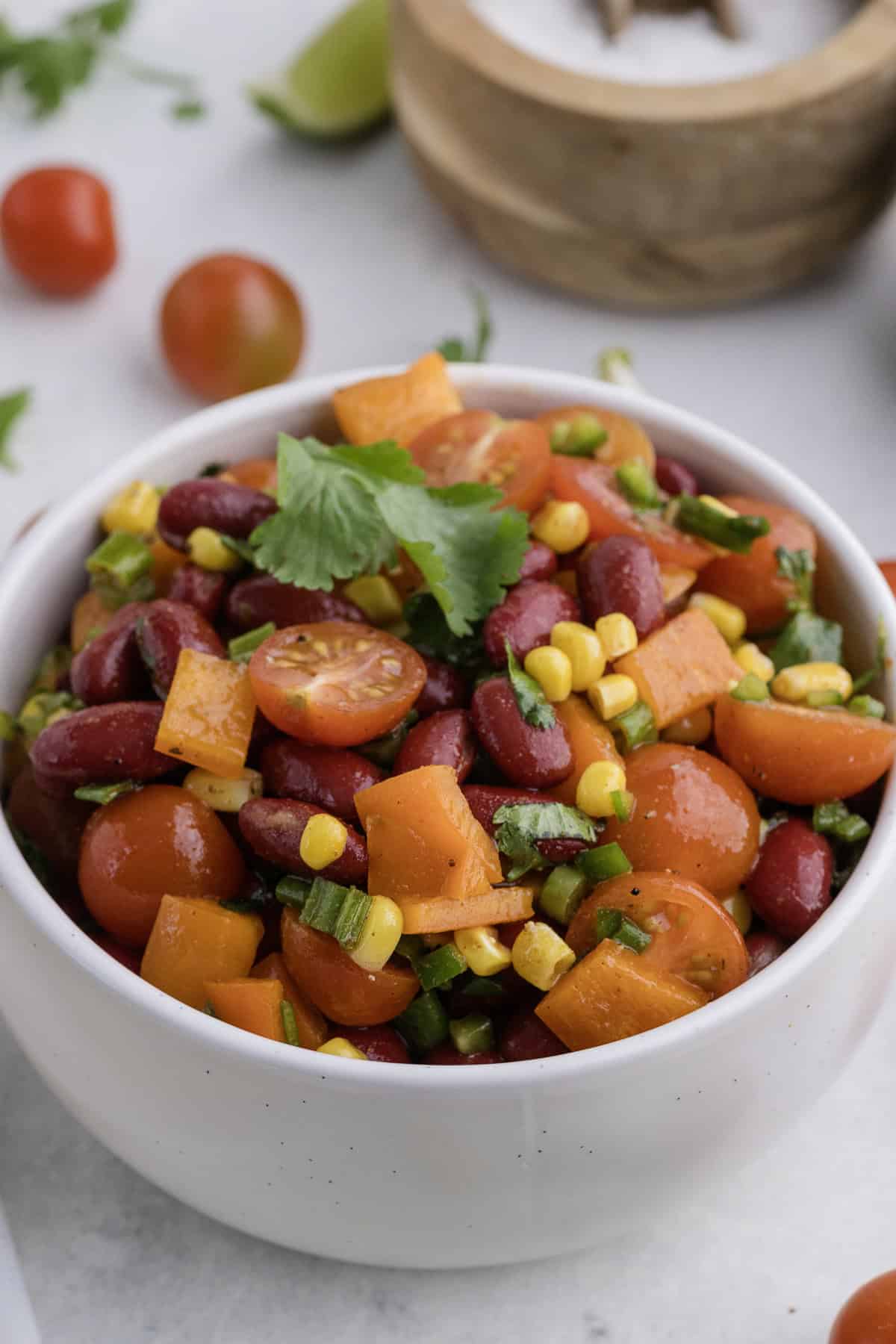 A white bowl filled with a colorful salad of kidney beans, cherry tomatoes, corn, bell peppers, and cilantro. There are tomatoes and a wooden bowl in the background.