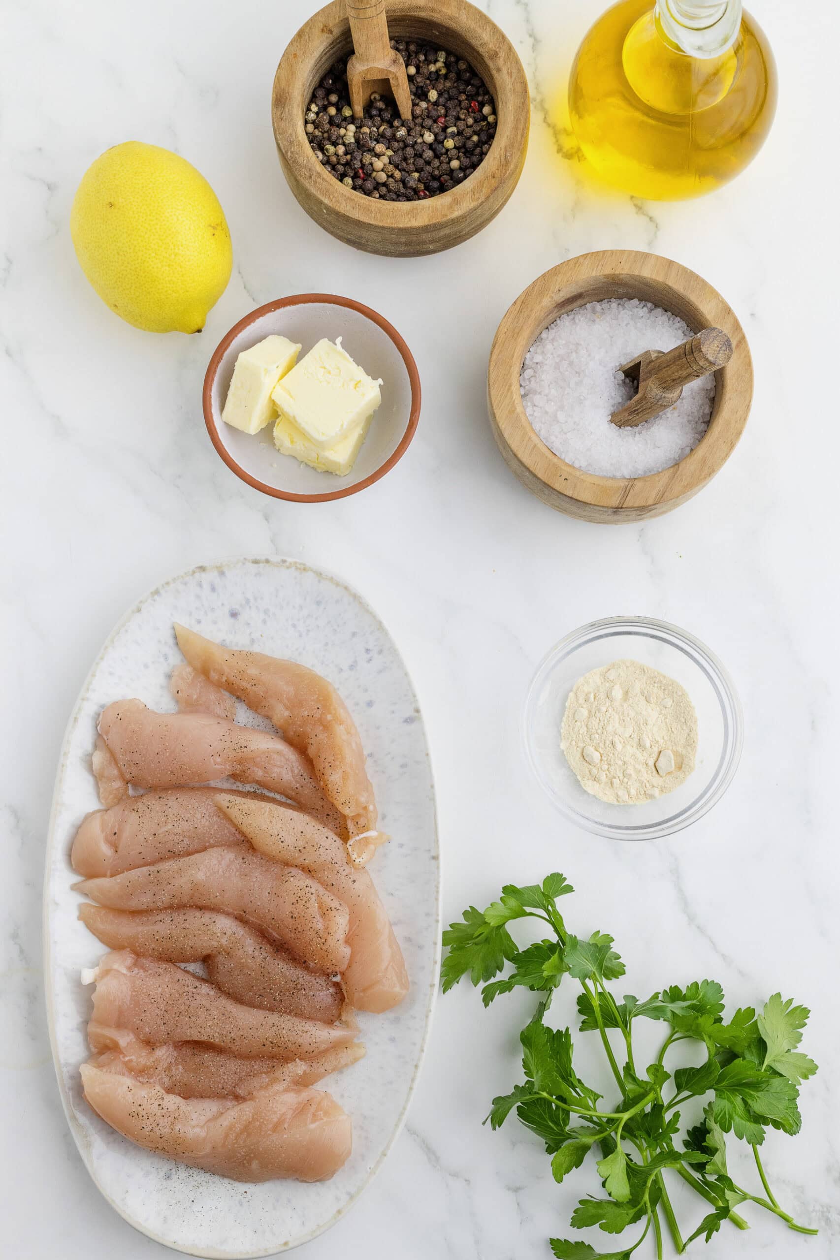 Chicken tenders on a plate with spices, lemon, butter, oil, and parsley on a marble surface.