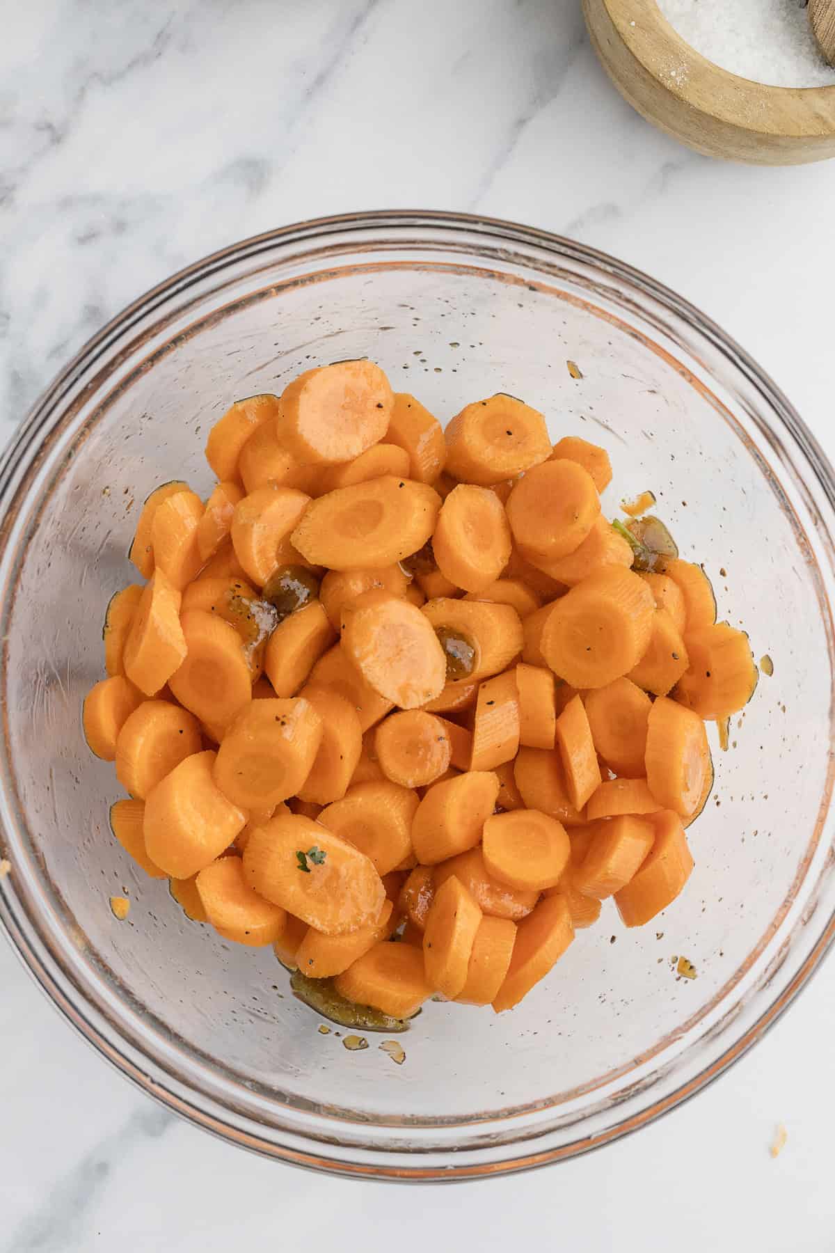 A glass bowl filled with sliced carrots mixed with oil and herbs. The bowl is placed on a marble countertop near a small dish of salt.