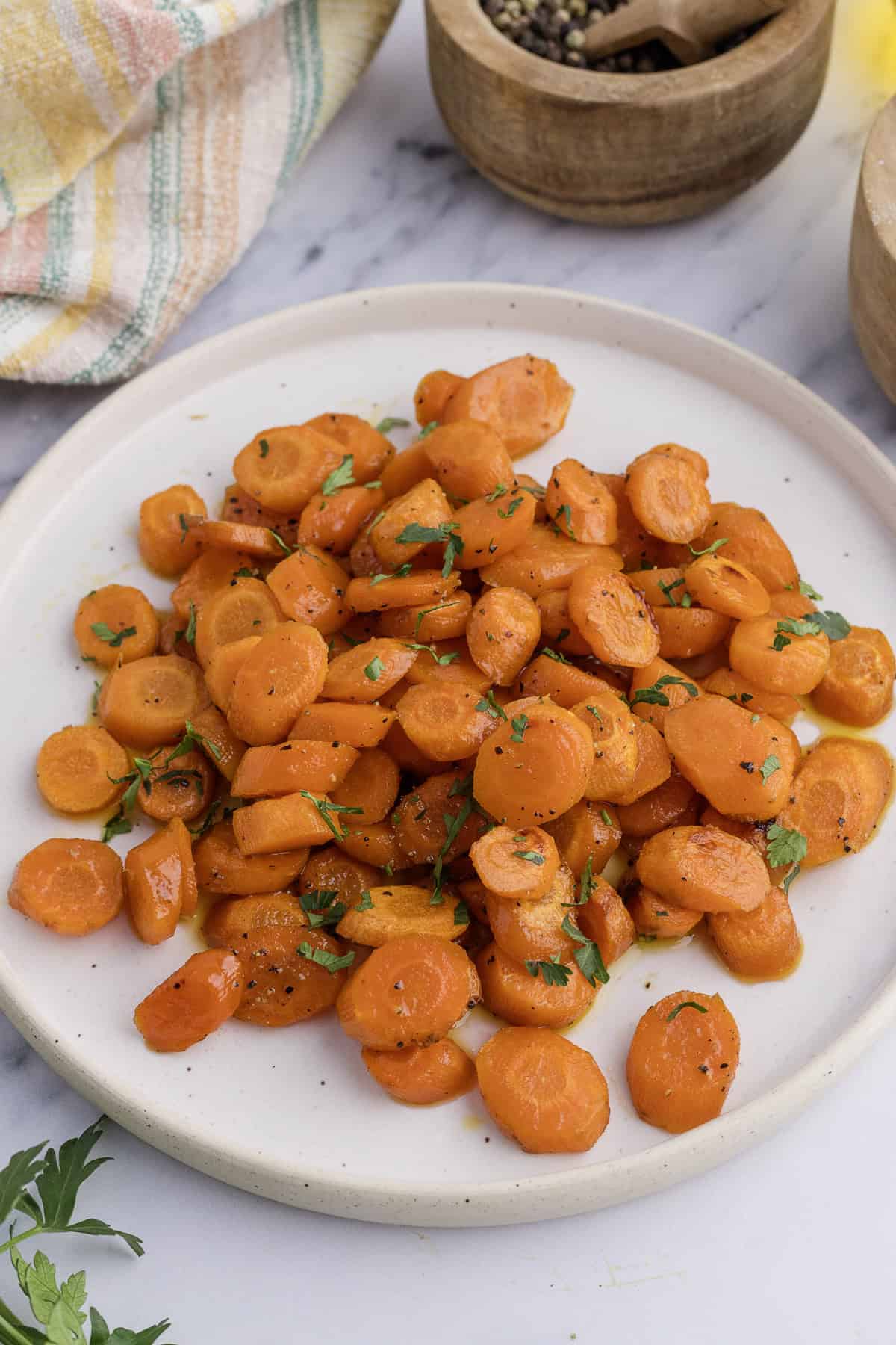 A plate of sliced, cooked carrots garnished with chopped herbs on a marble surface. A striped cloth and wooden bowls are in the background.