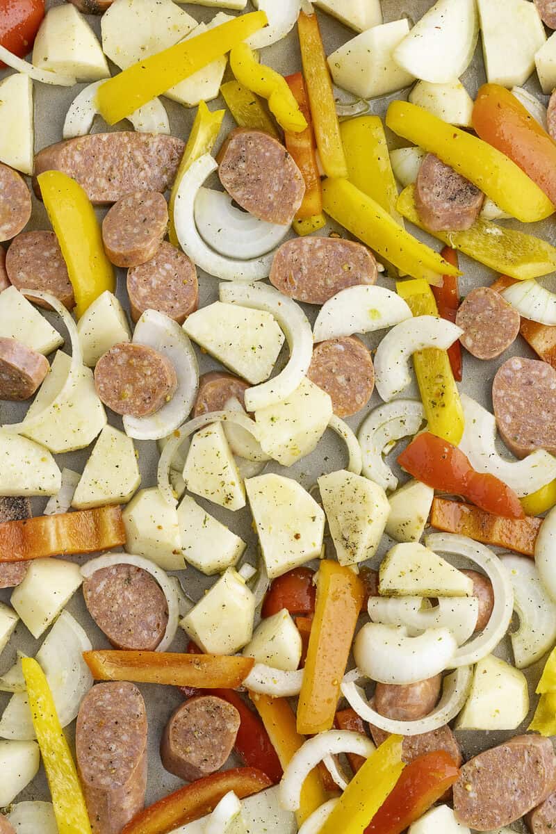 A sheet pan with sliced sausages, potatoes, onions, and bell peppers. The ingredients are seasoned and ready for roasting.