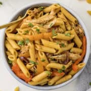 A bowl of penne pasta with shredded chicken, sliced bell peppers, red onions, and garnished with chopped parsley.