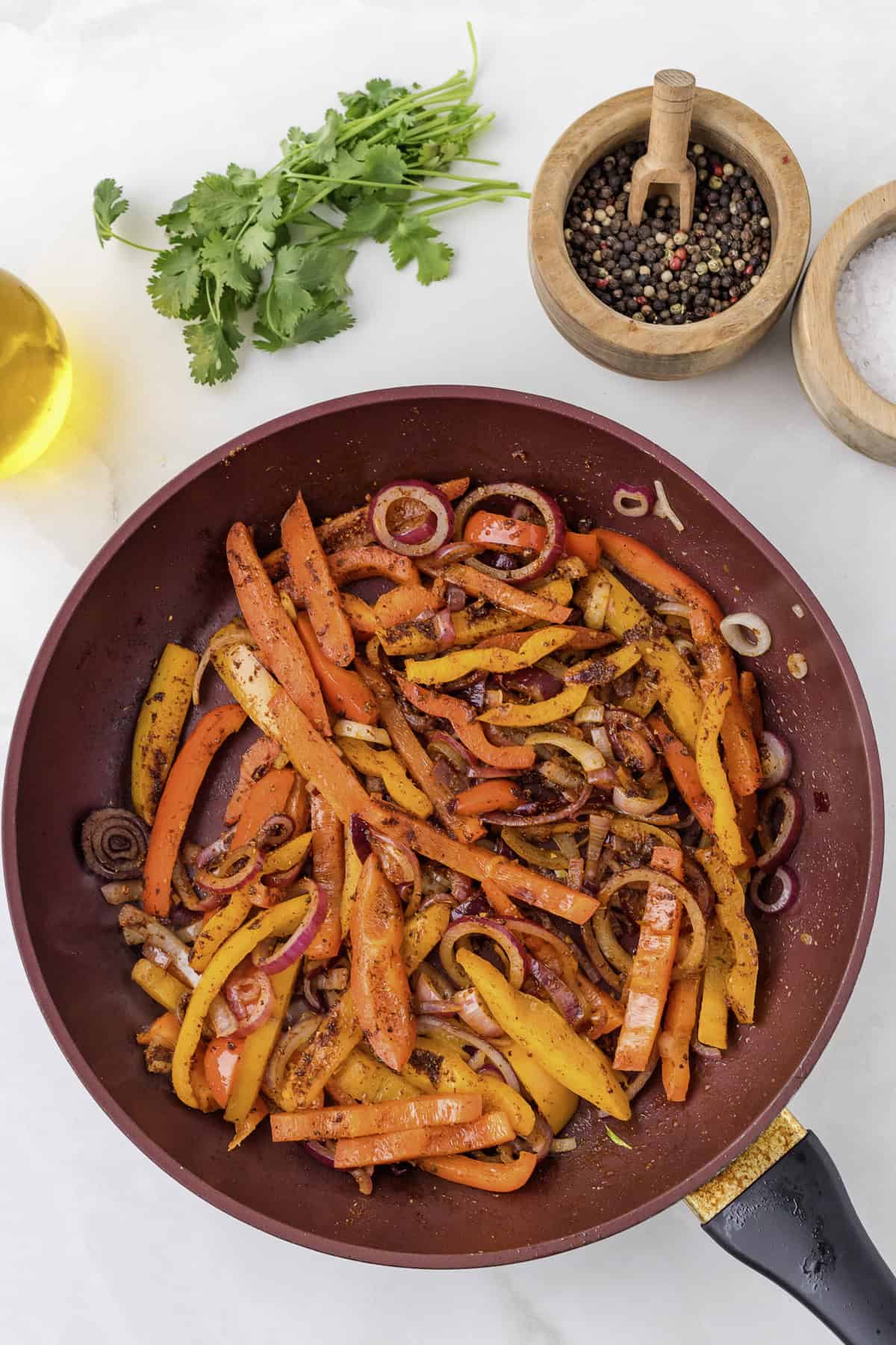 Sautéed red and yellow bell peppers with onions in a frying pan. Salt, pepper, oil, and cilantro are placed nearby on a white surface.