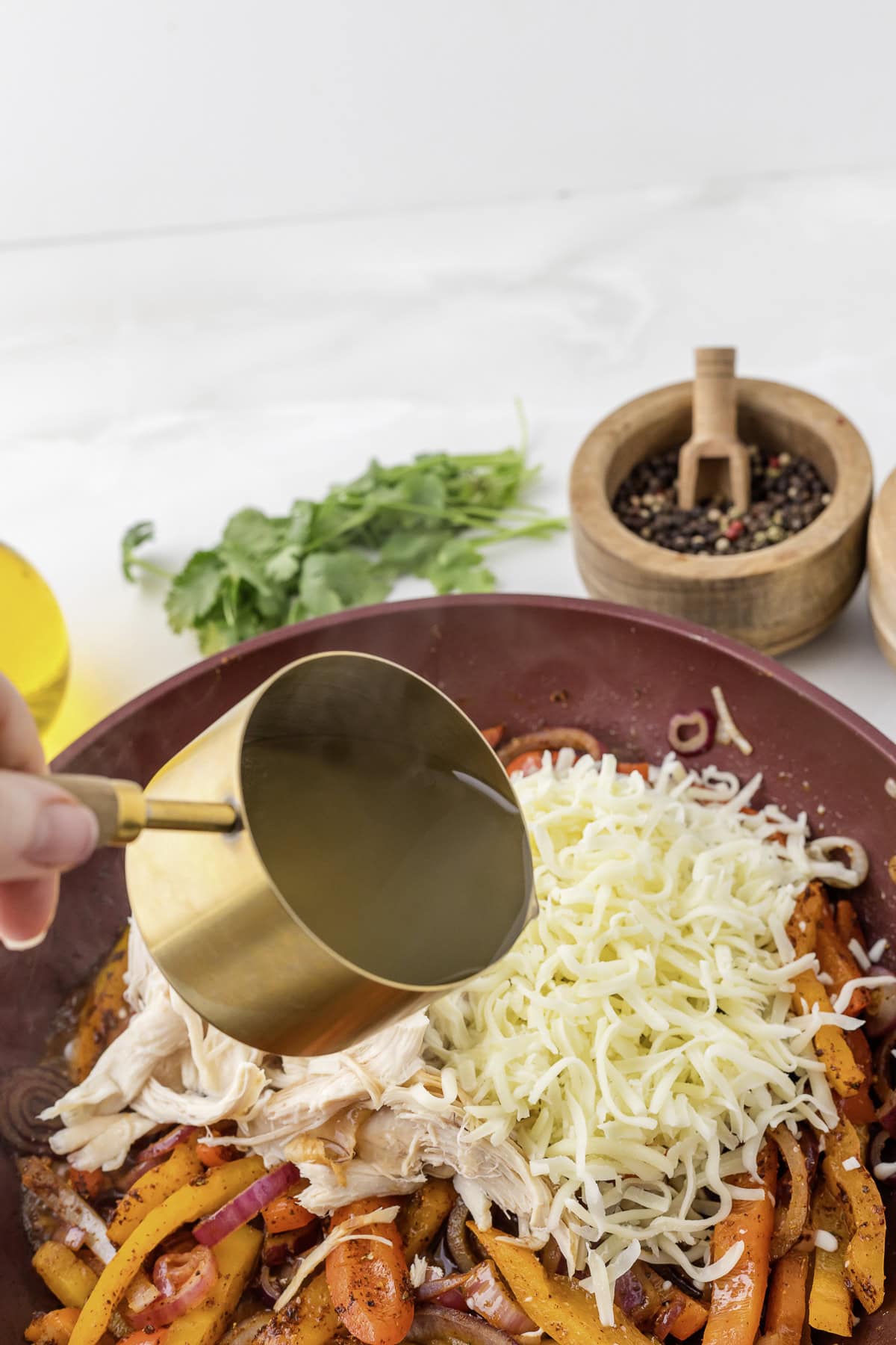 A cup of liquid is being poured over a skillet containing shredded cheese, cooked vegetables, and chicken. Nearby are a bowl of peppercorns and fresh herbs.